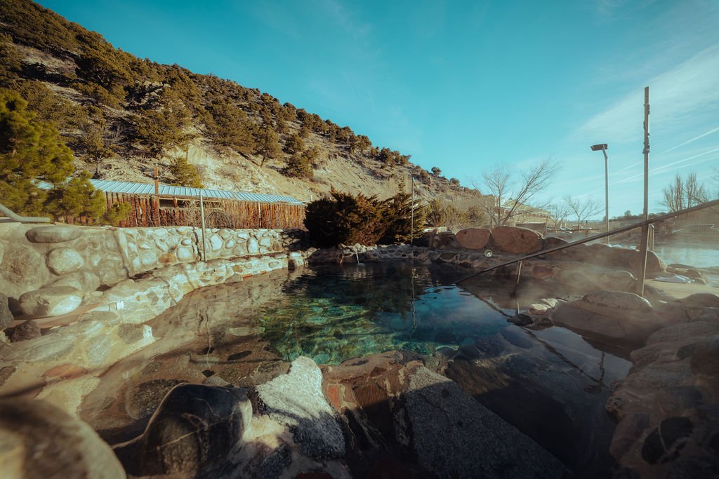 Outdoor hot spring or pool with rocks, stone wall, and mountain landscape in the background under a blue sky.