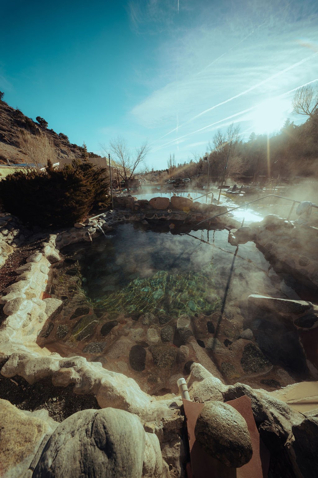 An outdoor hot spring pool with steam rising, surrounded by rocks and dry vegetation, with the sun shining brightly in a clear blue sky.