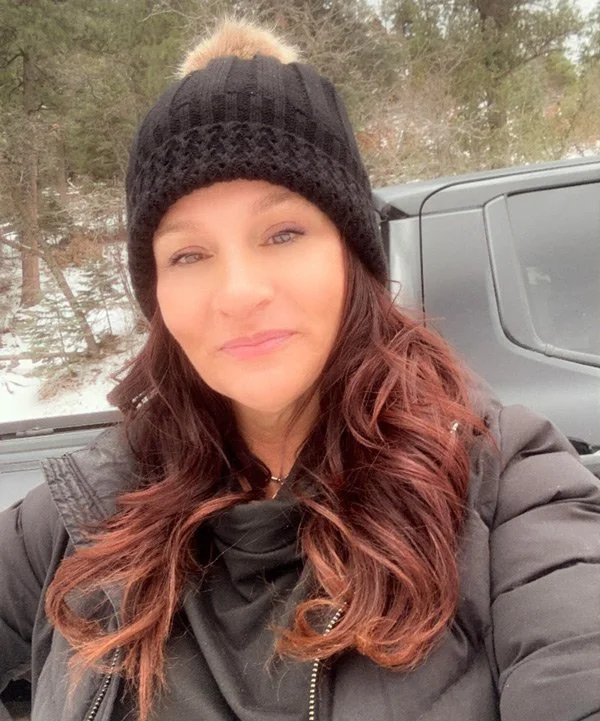 Woman with long wavy auburn hair taking a selfie outdoors in winter, wearing a black knit hat and black puffy jacket, with trees and a vehicle in the background.