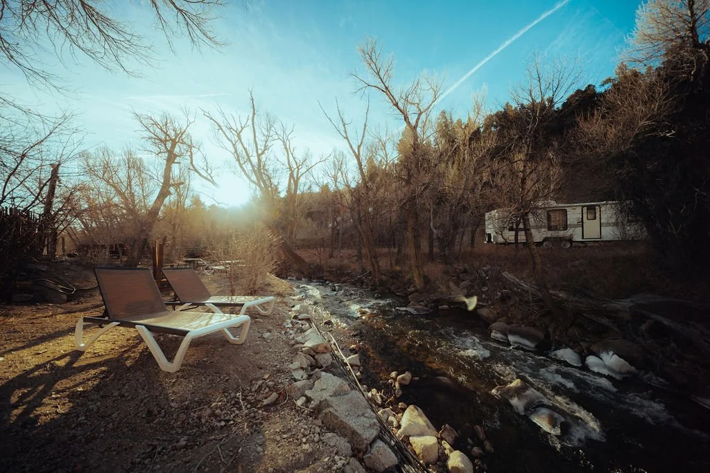 Sunset over a small stream with two lawn chairs on the rocky bank, leafless trees, and an RV trailer parked on a hillside, with a clear blue sky and a contrail.