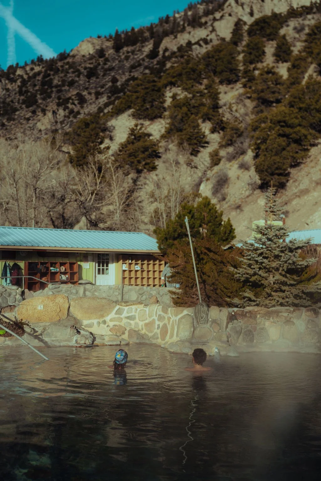 Two people swimming in an outdoor hot spring pool surrounded by rocks with trees, a building, and mountain landscape in the background.