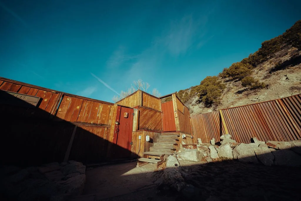 A rustic backyard with a wooden gate, stairs, and fencing, set against a hillside with trees under a clear blue sky.