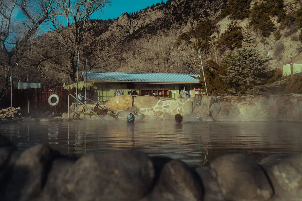 People enjoying a hot spring pool outdoors with steam rising, surrounded by rocks, trees, and a building with a blue roof in a mountainous area.