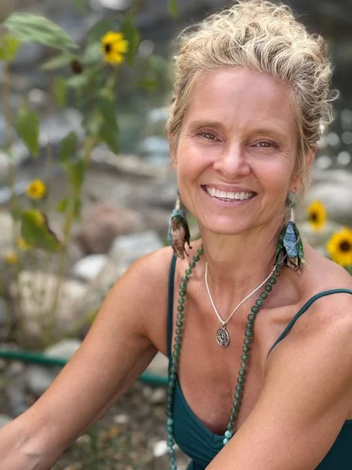 A smiling middle-aged woman with short, curly blonde hair wearing earrings, a necklace, and a teal spaghetti strap top, outdoors with yellow flowers and greenery in the background.