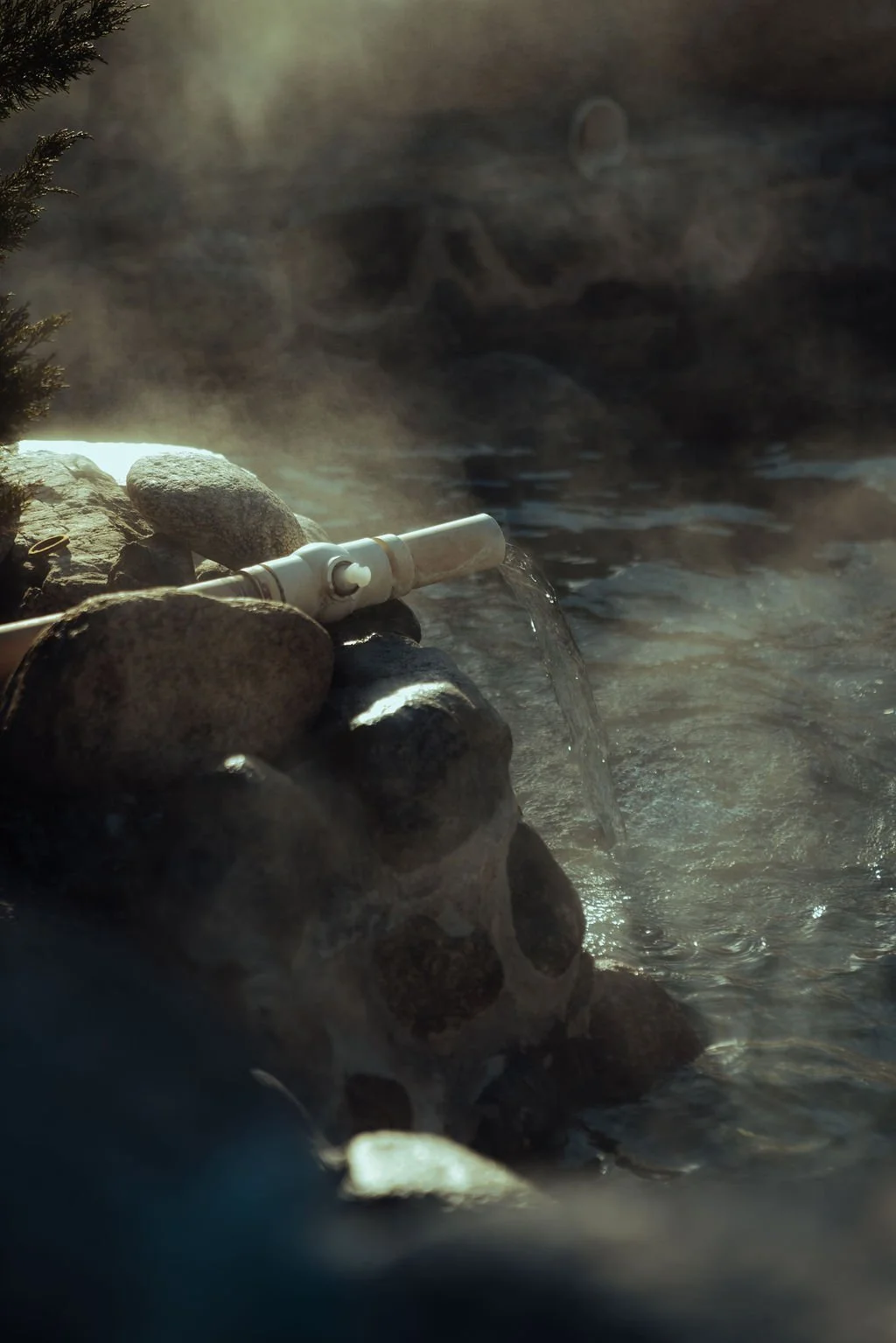 A PVC pipe positioned among rocks releasing water into a hot spring, with steam rising in the background.