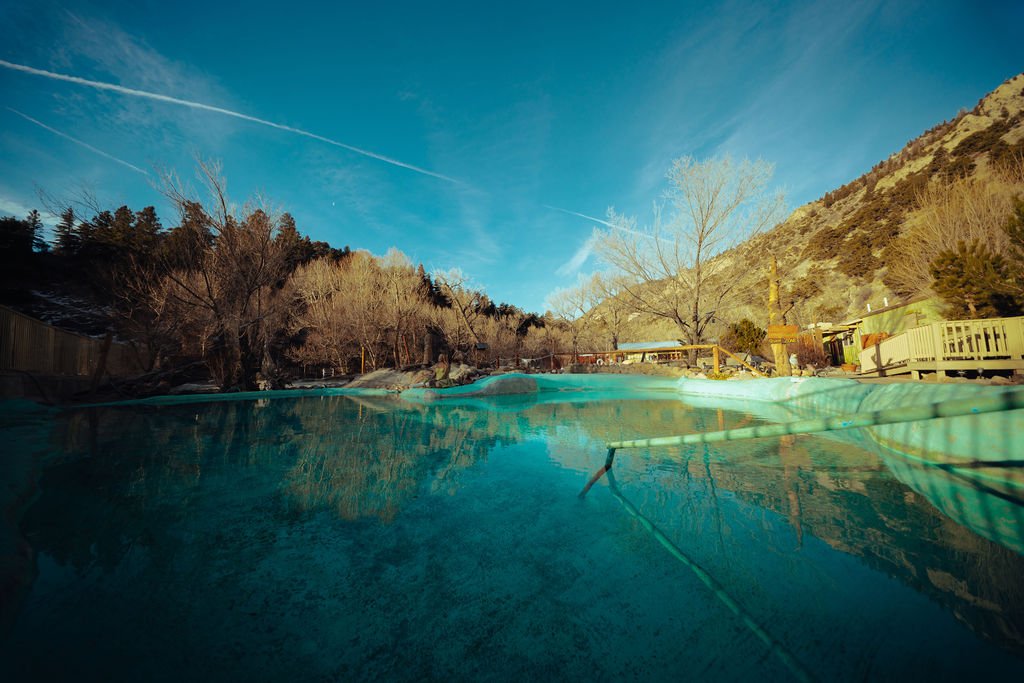 A natural outdoor hot spring pool in a mountainous area during winter, with leafless trees, a wooden deck, and a clear blue sky.