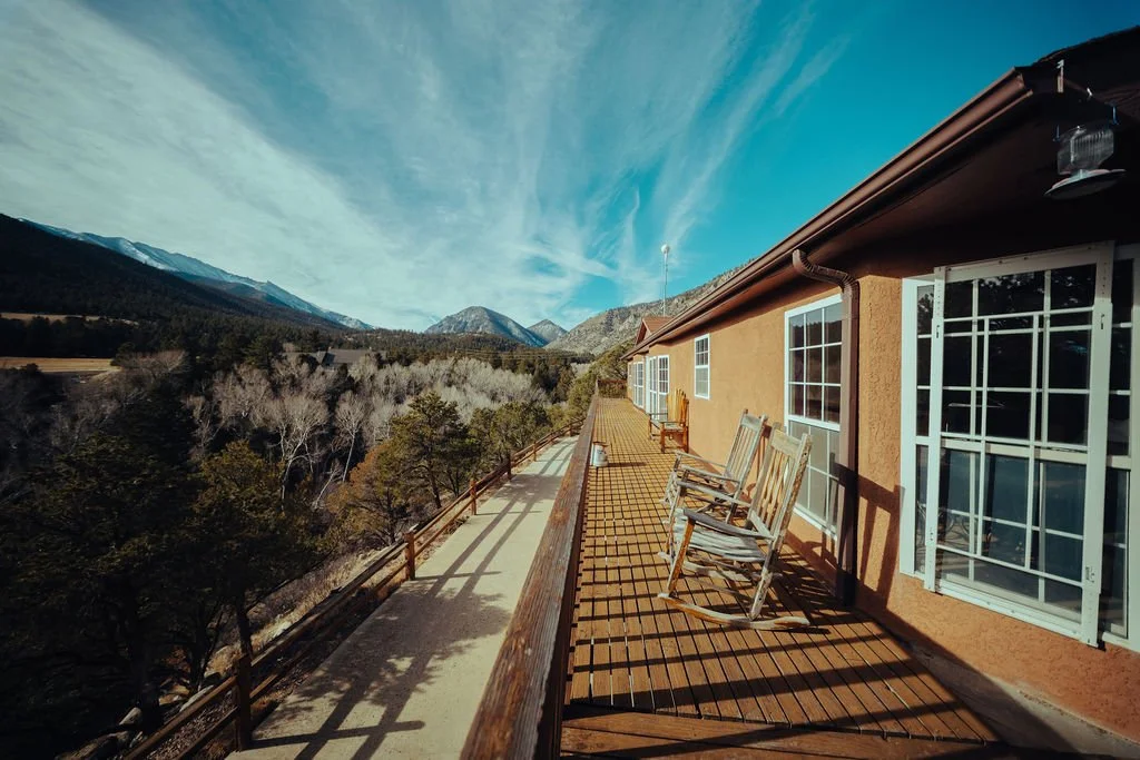 A balcony with wooden chairs on an orange building, overlooking a forested valley and mountains under a blue sky.