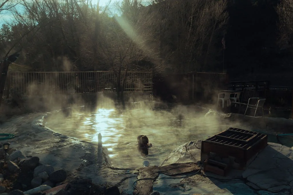 A person floating in an outdoor hot spring or spa pool surrounded by steam during late afternoon or early evening, with chairs and railing in the background.