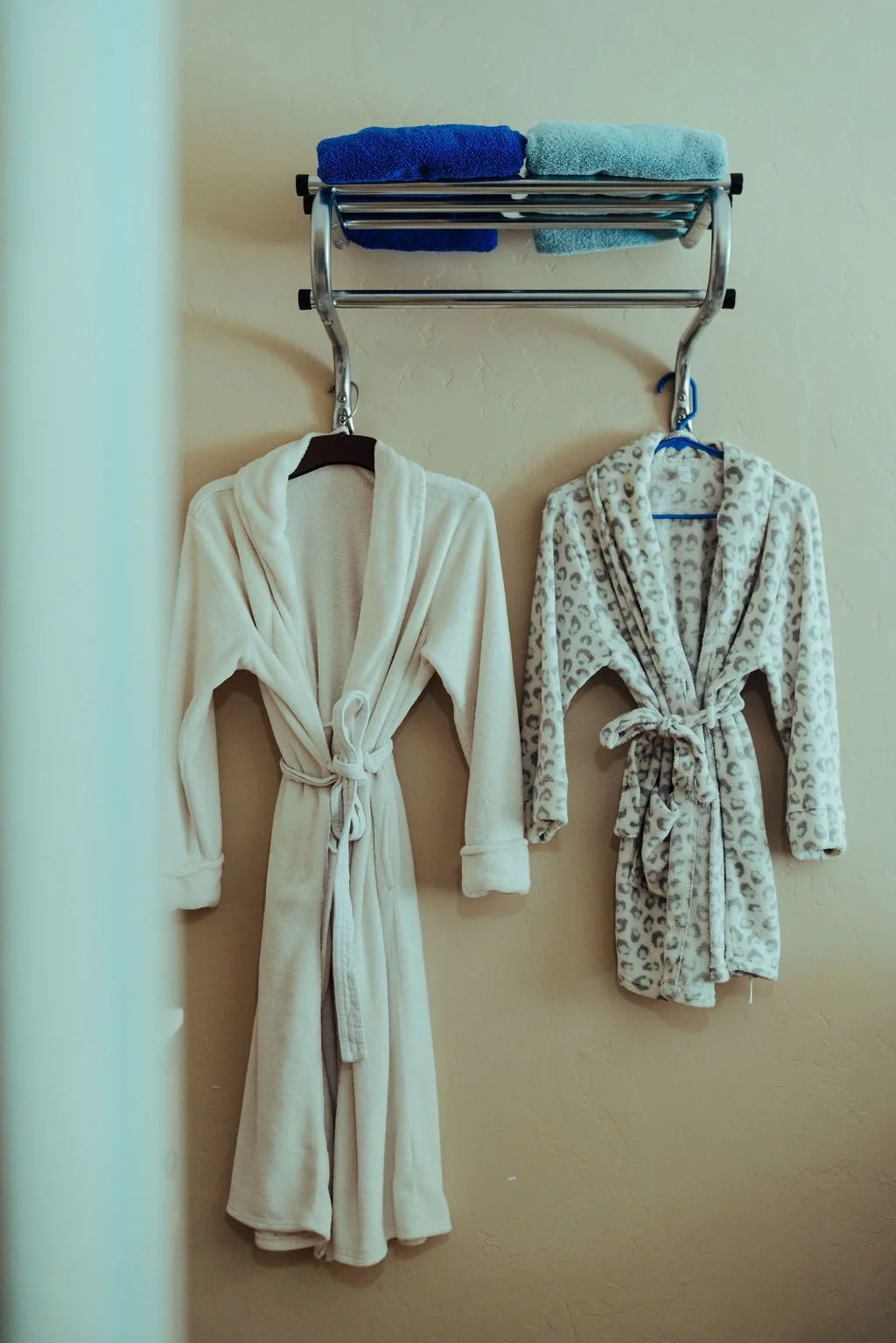 Two bathrobes hanging on a wall, one cream-colored and one animal print, with towels on a shelf above.
