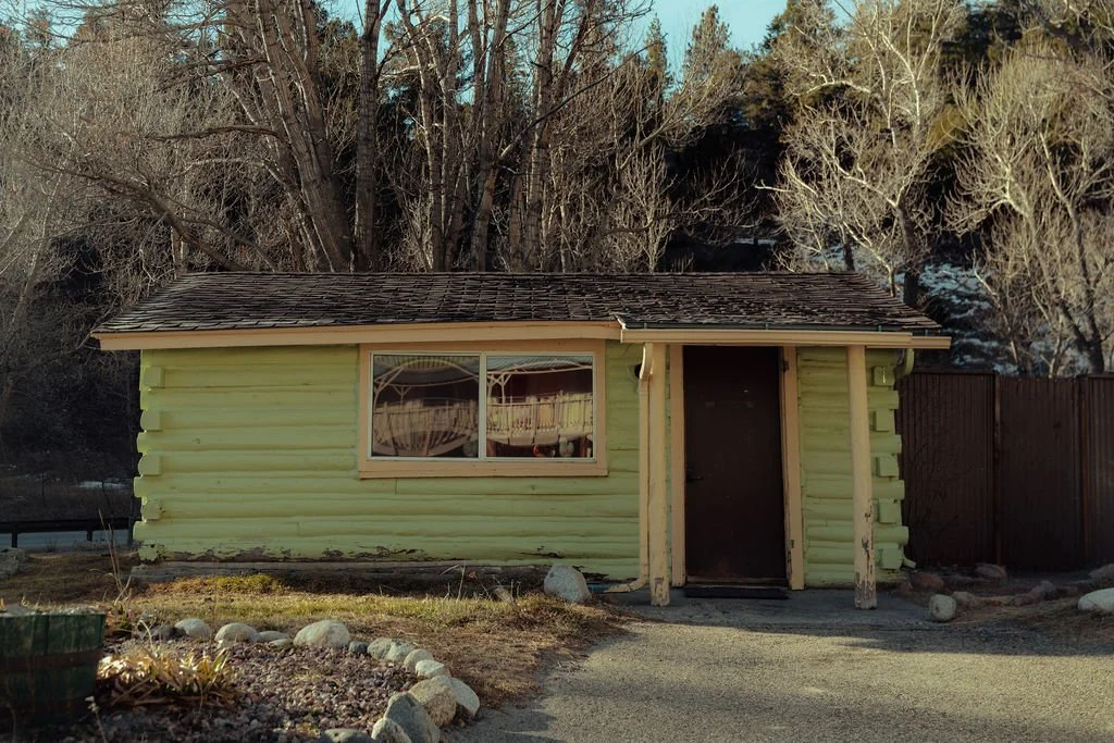 A small yellow wooden building with a brown shingled roof, a single window, and a door, located outdoors with leafless trees in the background.