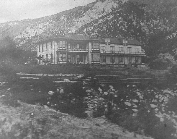 Black and white photo of a large wooden house or lodge in a mountainous area, with rocky terrain and cliffs in the background.