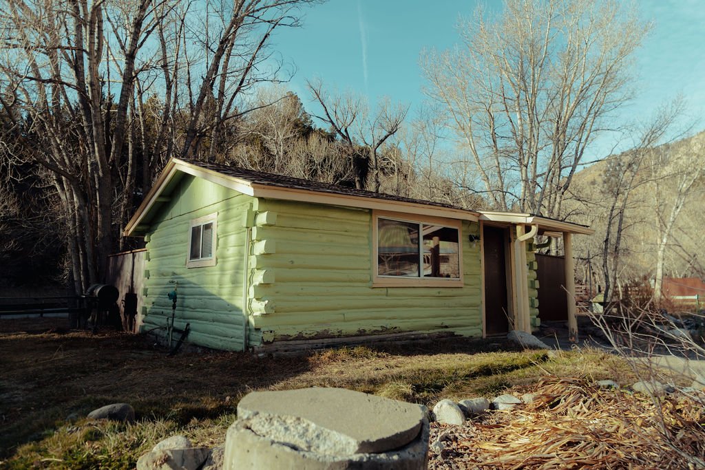 A small, single-story house with light green wooden siding and white trim, situated on a grassy yard with some rocks and fallen leaves, surrounded by leafless trees on a sunny day.