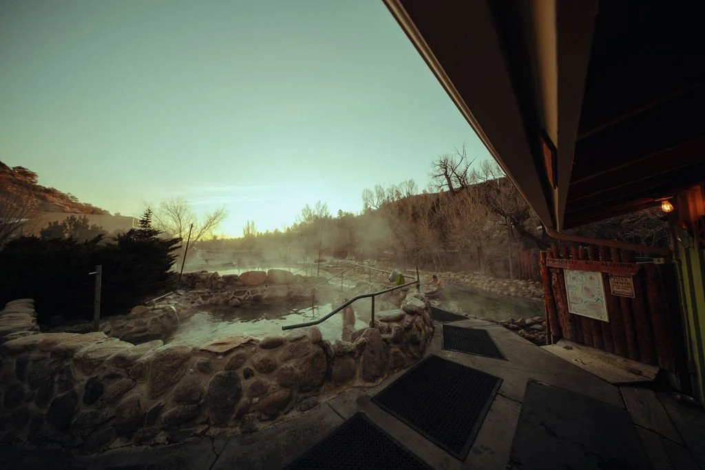 Outdoor hot spring with rocky edge and steam rising, surrounded by trees under a greenish sky, with wooden structure on the right.