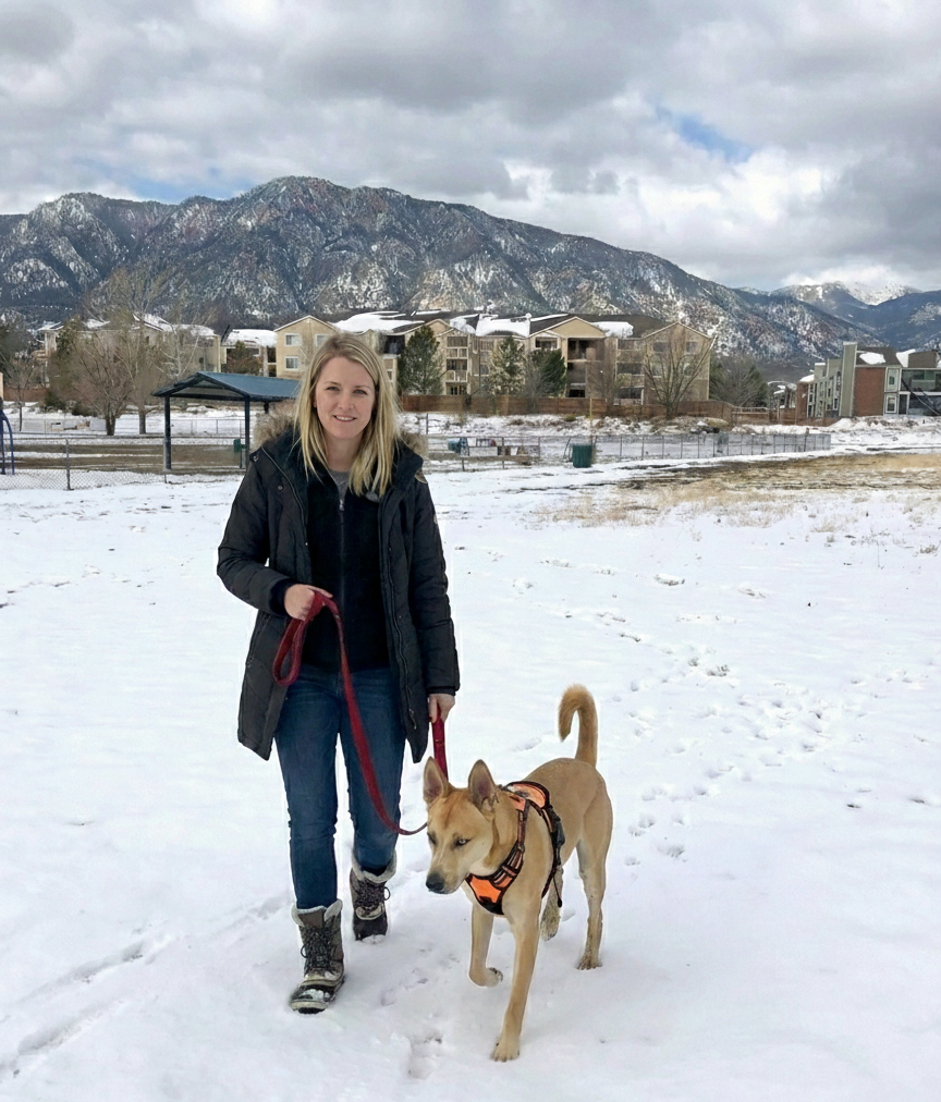 A woman walking her dog in a snowy park with mountains in the background.
