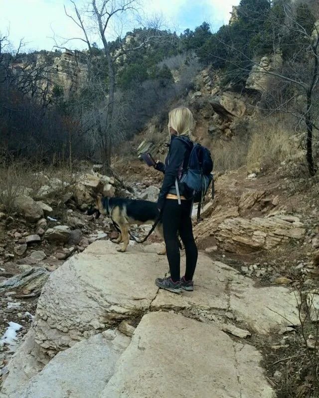 A woman with blonde hair in a ponytail, wearing a black jacket and black pants, is standing on a large rock in a mountainous area with trees and rocks, holding a phone, with a black and tan dog on a leash beside her.