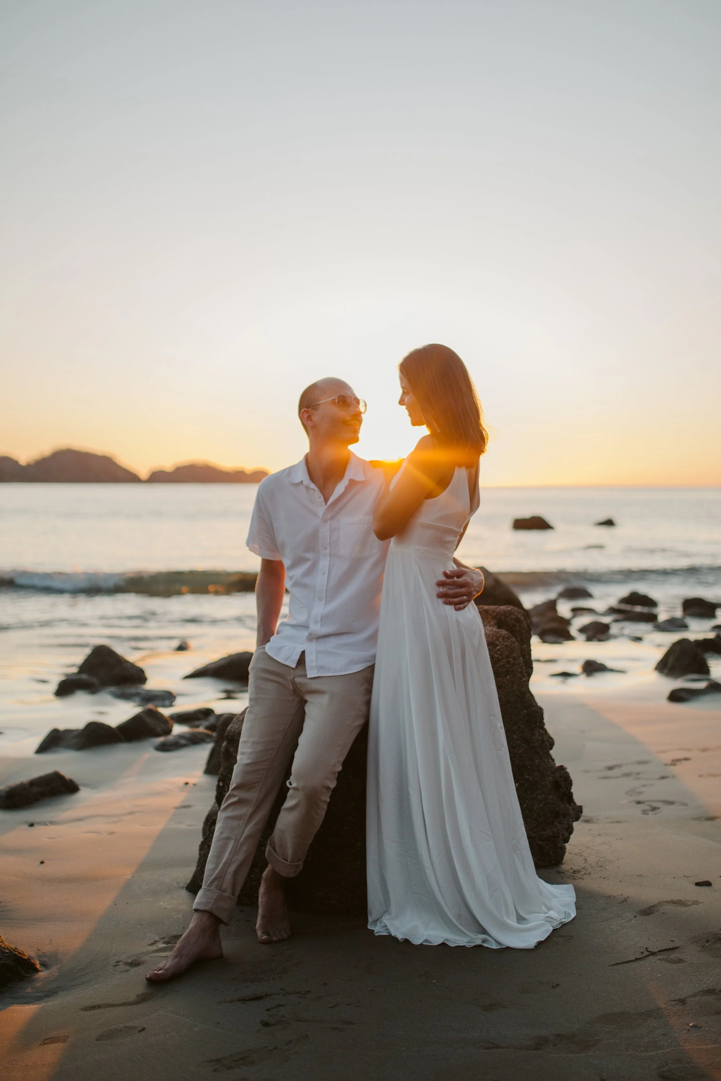 A couple standing on a beach at sunset; the woman in a white dress and the man in light-colored pants and a white shirt.