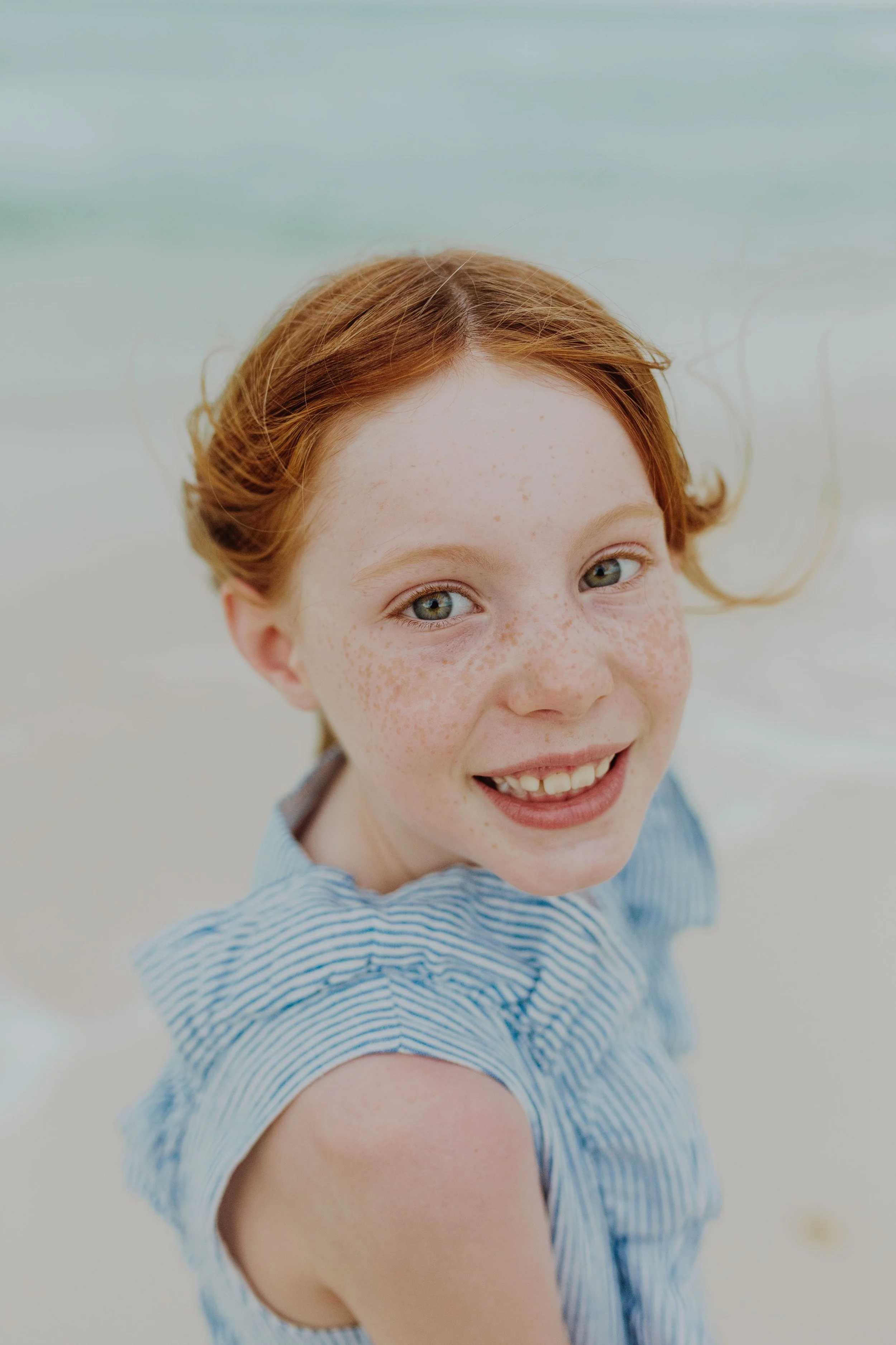 Close-up of a smiling young girl with red hair, freckles, and blue eyes, wearing a blue and white striped sleeveless top, outdoors with a blurred beach background.