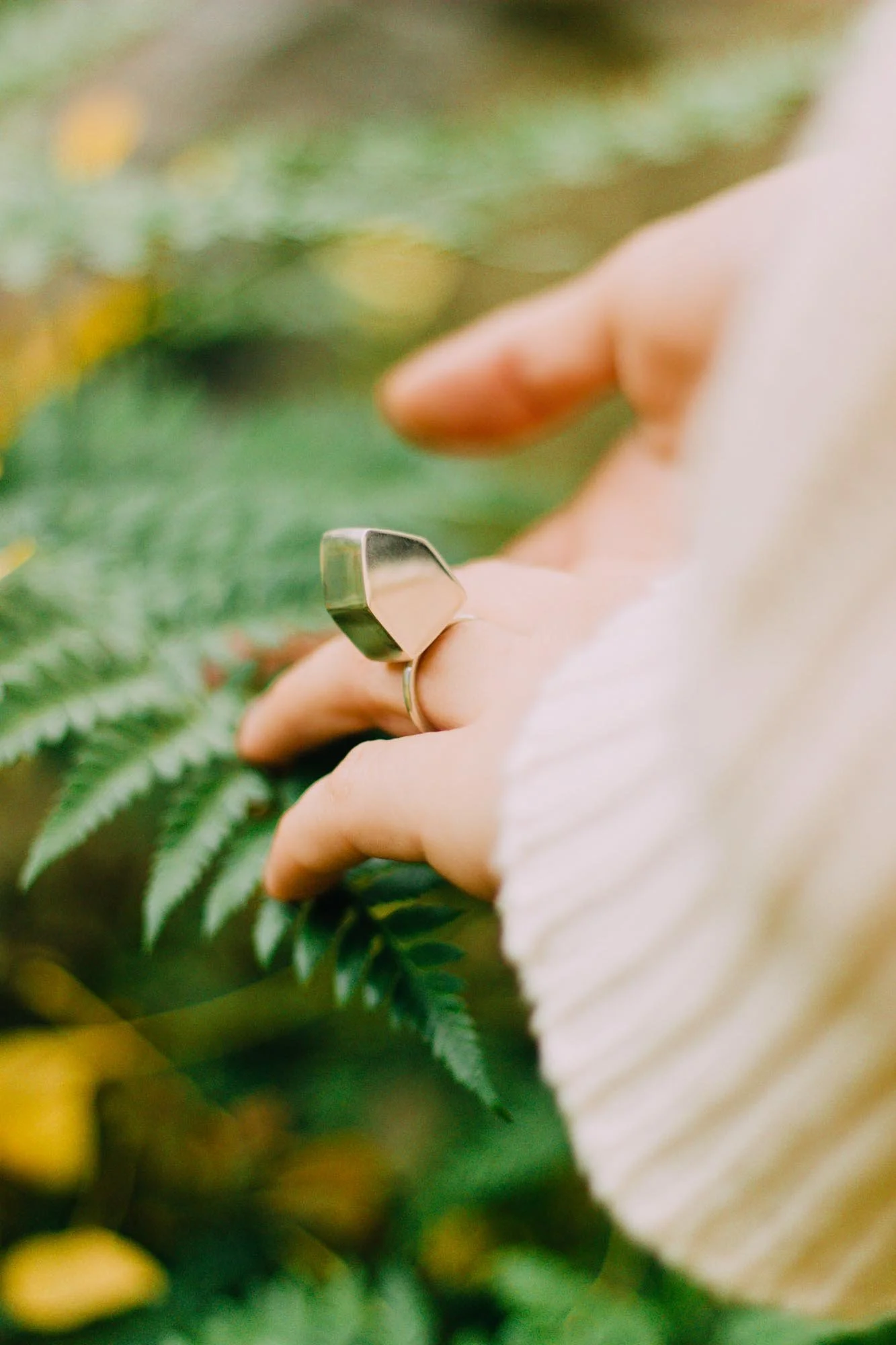 A person wearing a white or cream-colored knitted sweater is holding a fern leaf and a metallic ring on their finger.