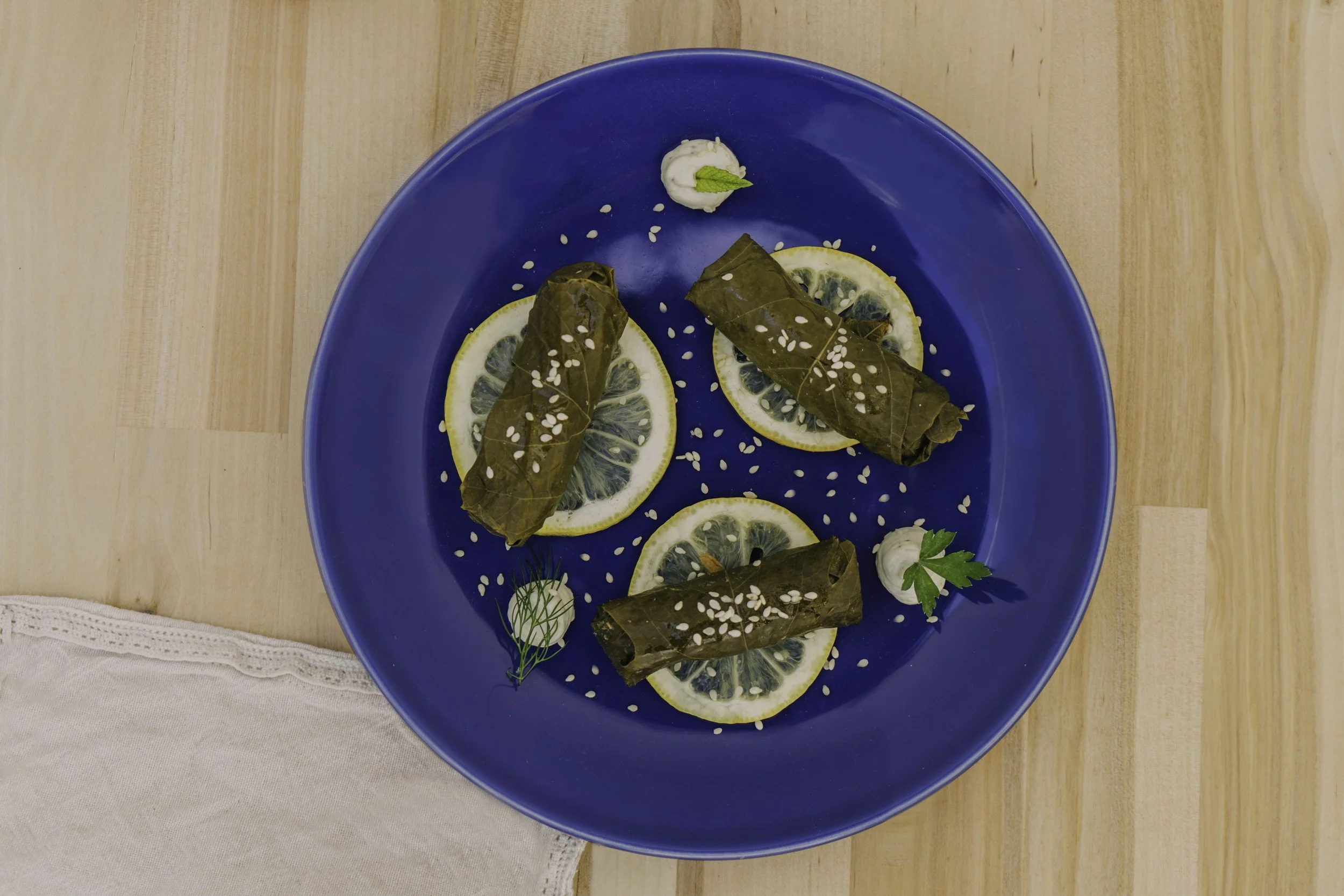 Plate with three stuffed grape leaves on lemon slices, garnished with sesame seeds, dill, parsley, and small dollops of white sauce on a wooden table.