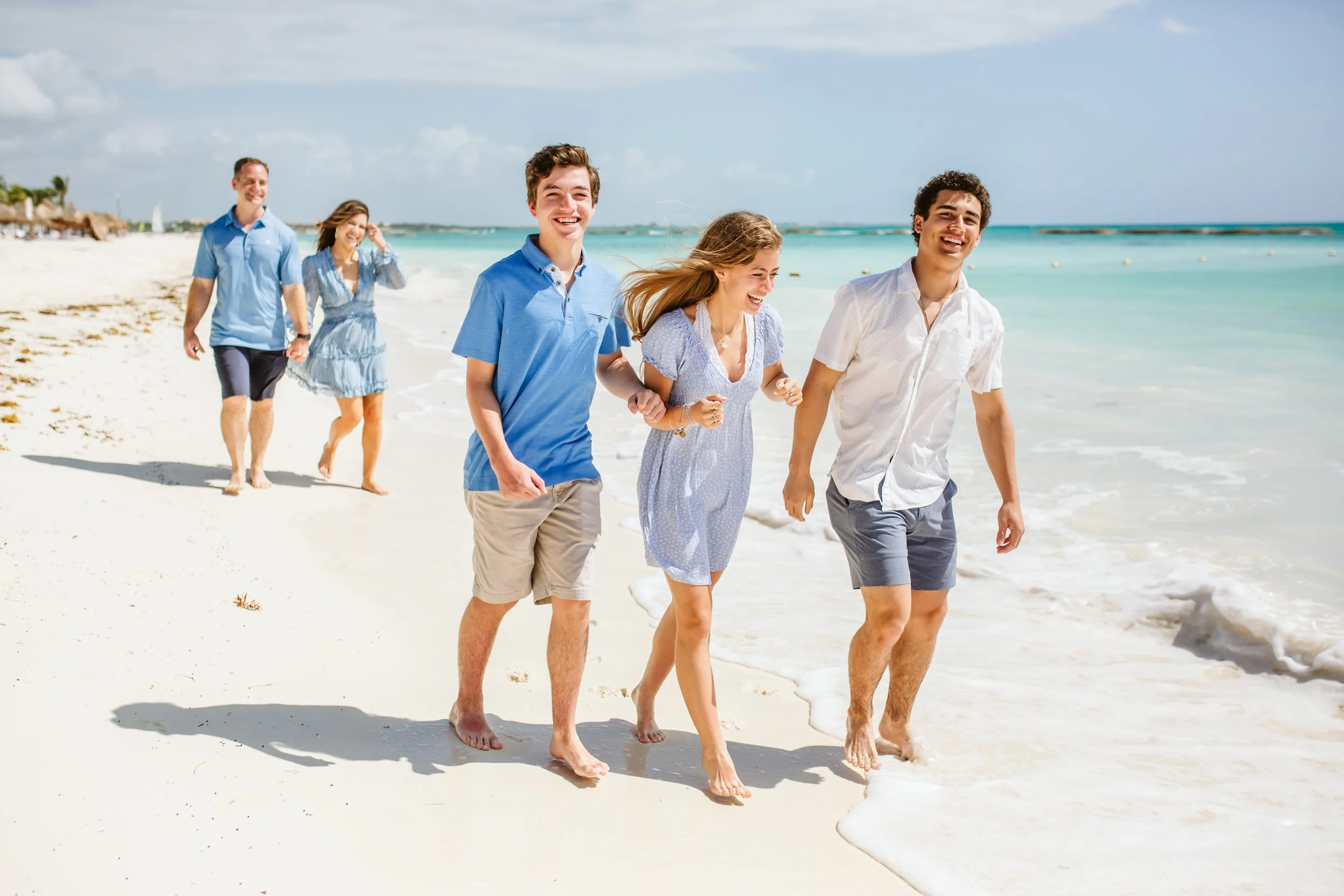 Five young adults walking along the beach near the shoreline, all smiling and enjoying the sunny day.