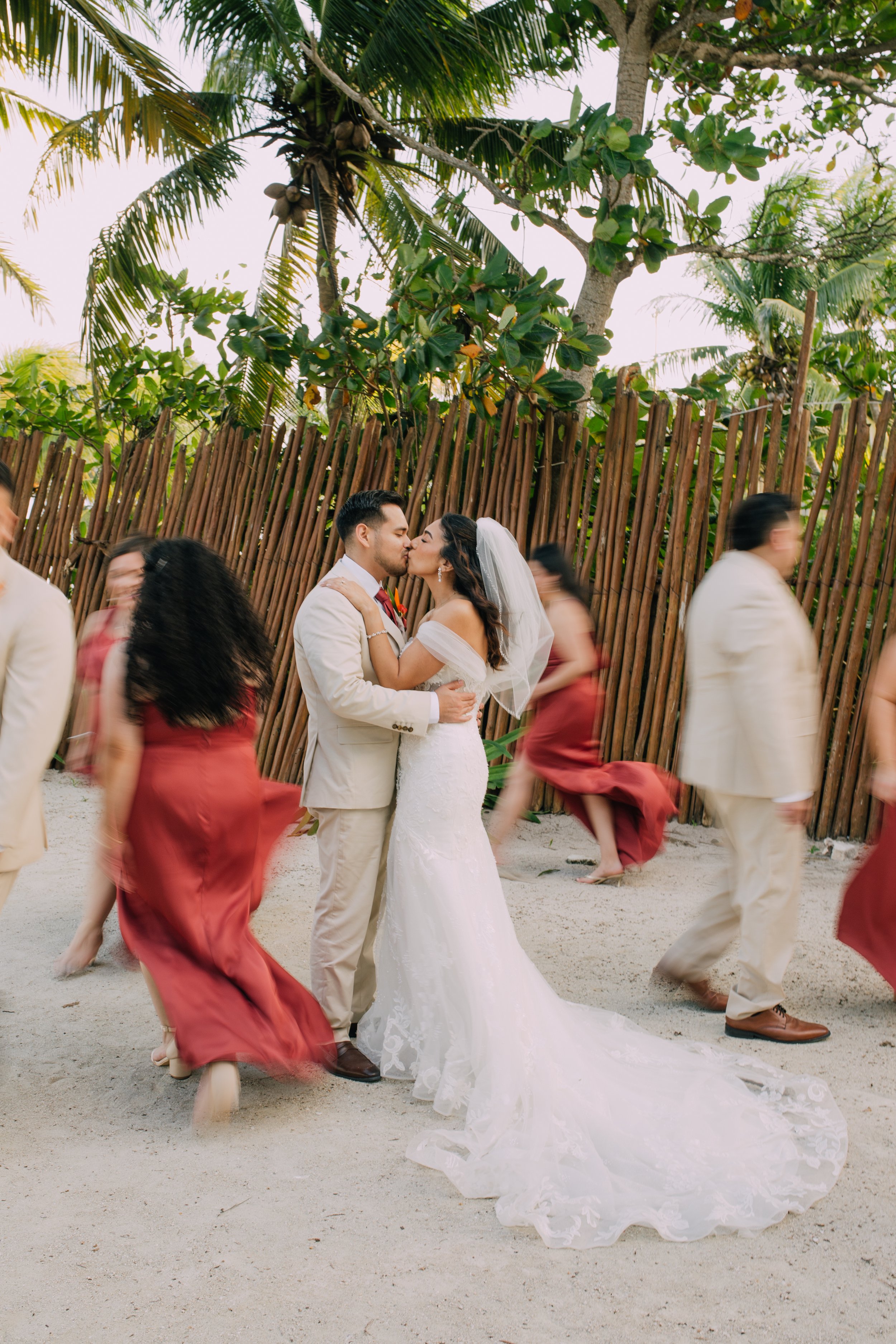 A bride and groom kissing at their wedding on a beach, surrounded by wedding guests dancing and celebrating.