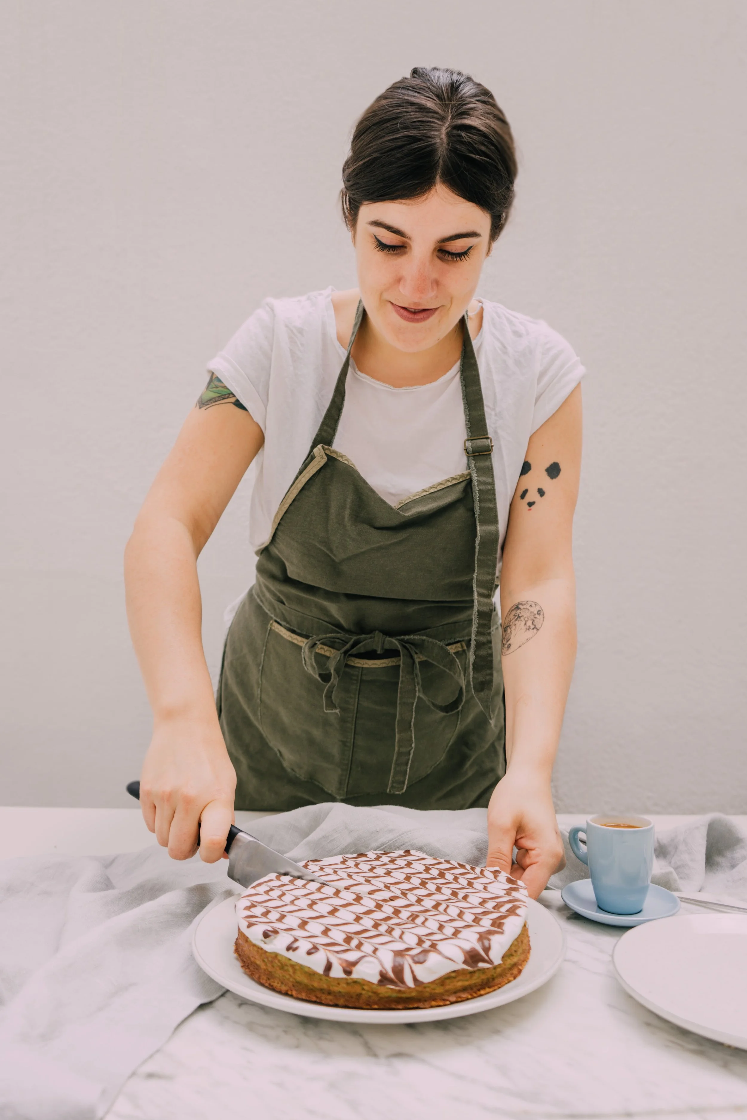 A woman in a green apron cuts a decorated cake with a knife. There is a cup of coffee on a saucer on the table beside her.