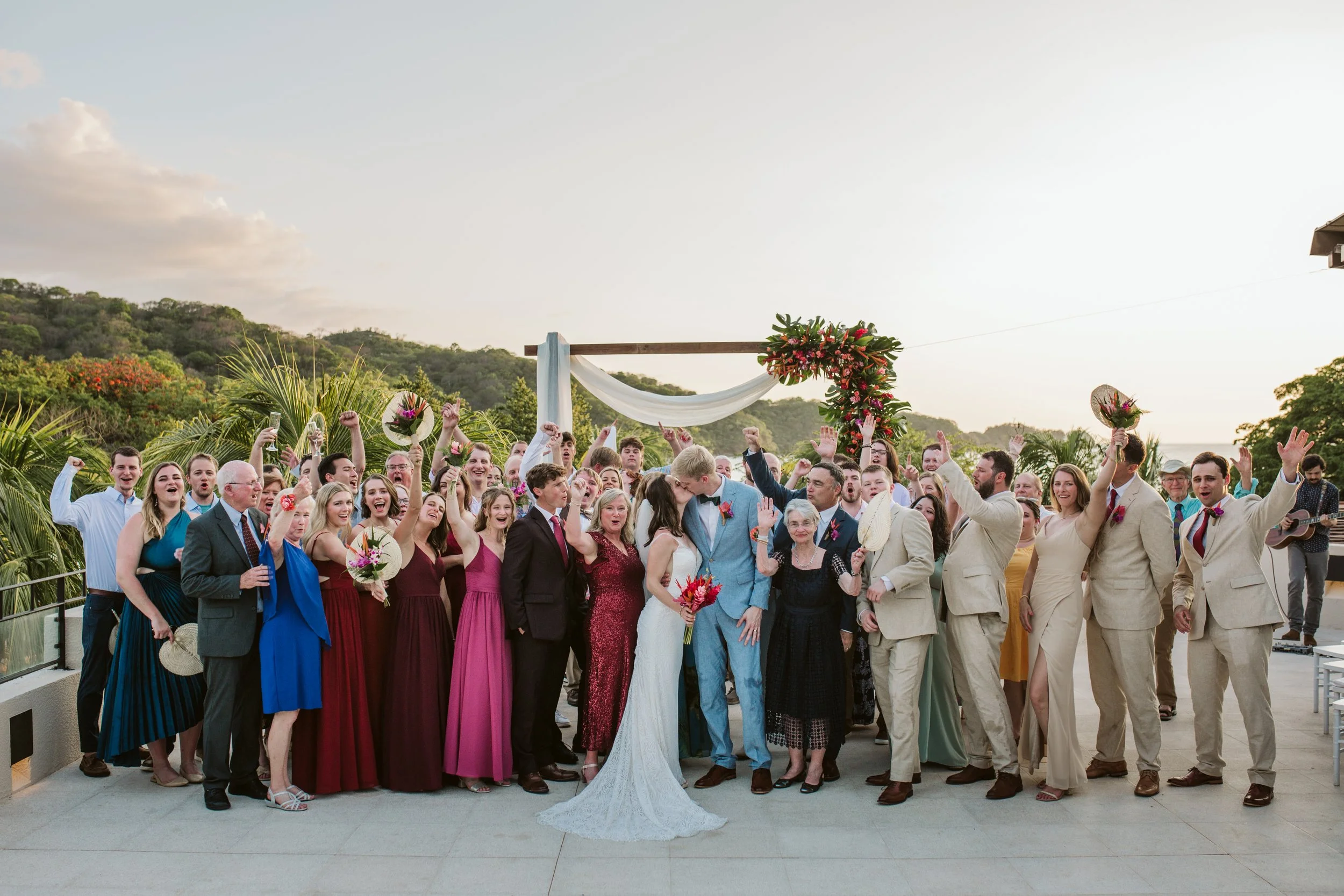 A large group of wedding guests and the bride and groom celebrate outdoors at sunset, with some holding bouquets and others raising their hands in celebration. The scene includes a scenic backdrop of green hills and a decorated arch.