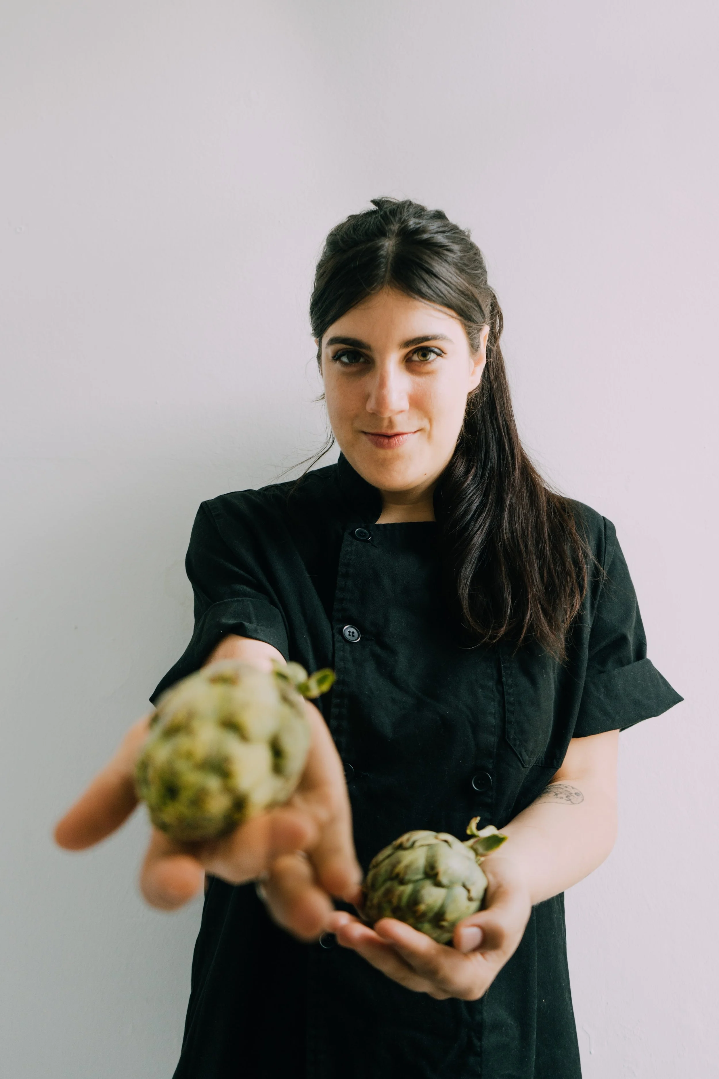 A woman with dark hair in a black chef's coat holding three artichokes, reaching towards the camera, against a plain light background.