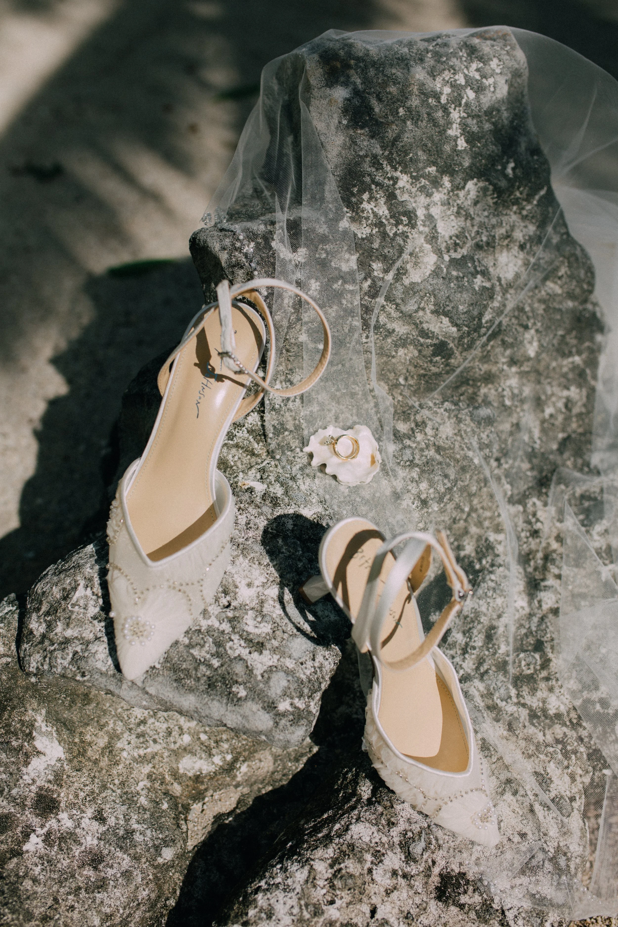 White bridal shoes with pearl decorations, a small white seashell ring holder with rings, and a delicate veil on large rocks outdoors.