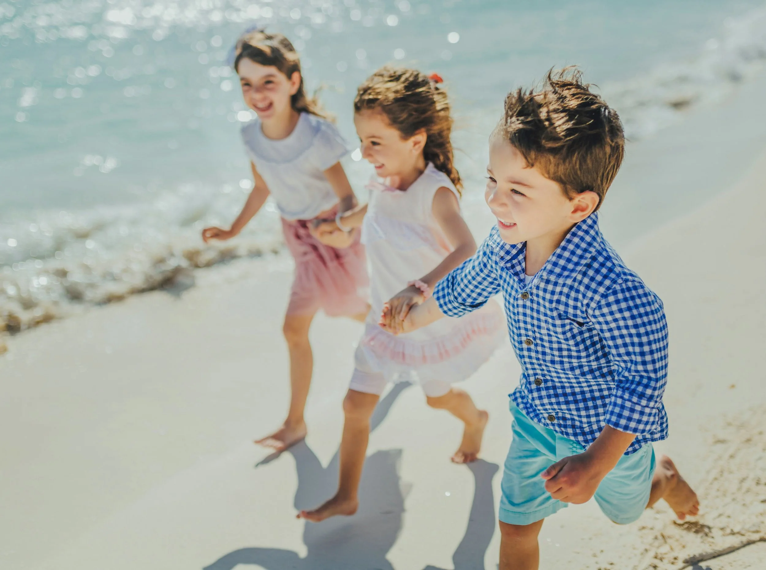 Four children playing and running hand-in-hand on a sunny beach with ocean waves in the background.