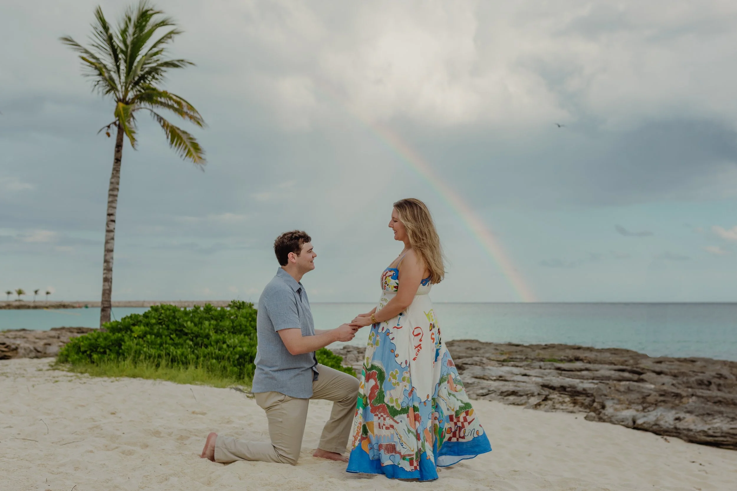 A man proposing to a woman on a beach with a rainbow in the sky, a palm tree, and the ocean in the background.