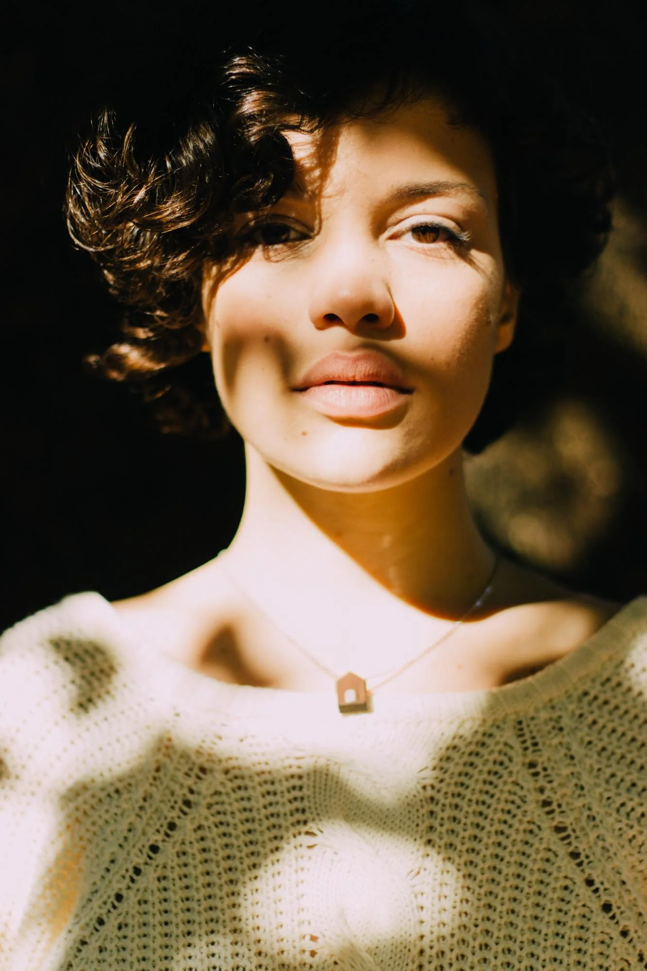 A woman with short curly hair wearing a cream knitted sweater and a necklace, standing outdoors in sunlight with shadows on her face.