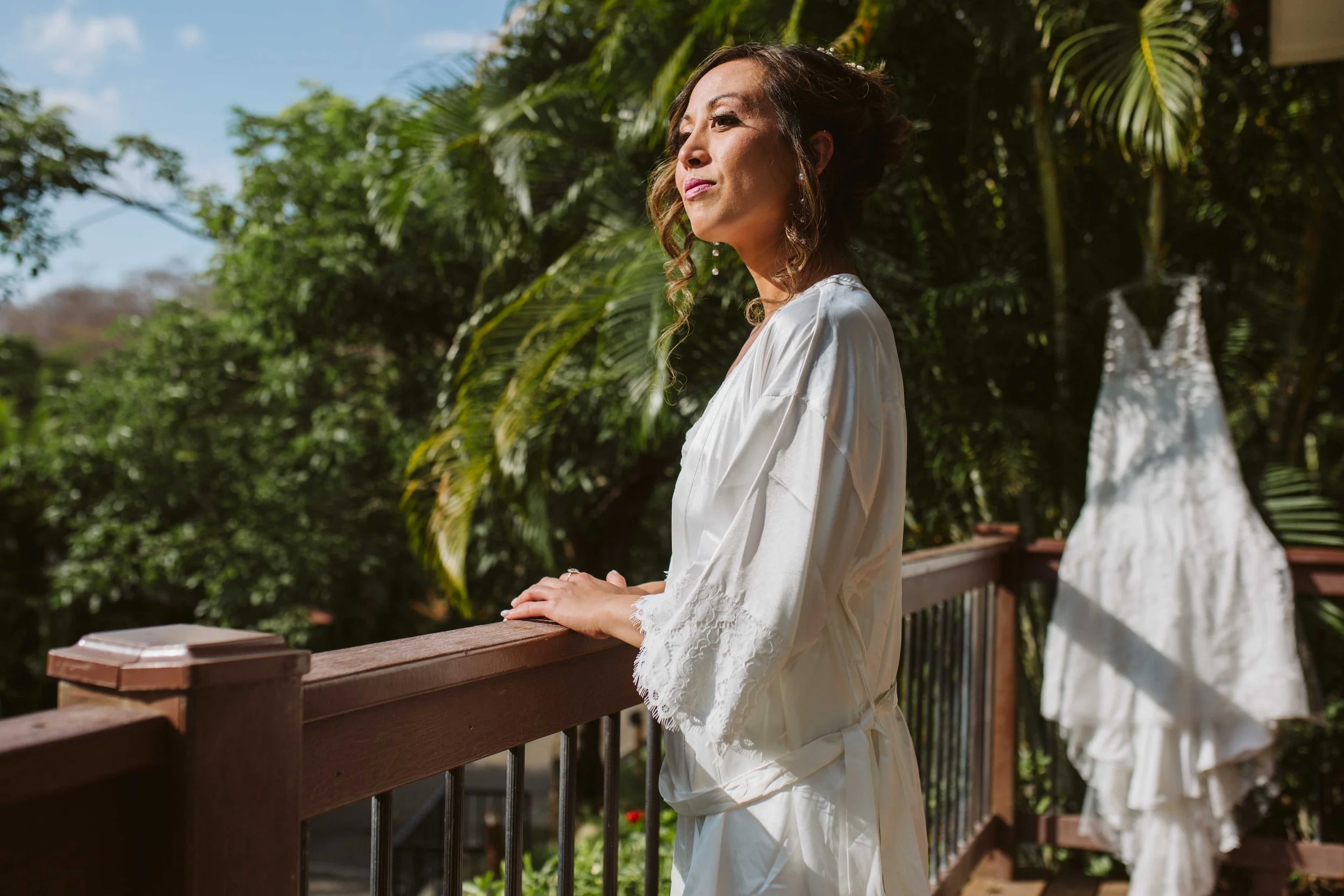 A woman in a white satin robe standing on a balcony with tropical trees in the background. A white wedding dress on a hanger is visible behind her.