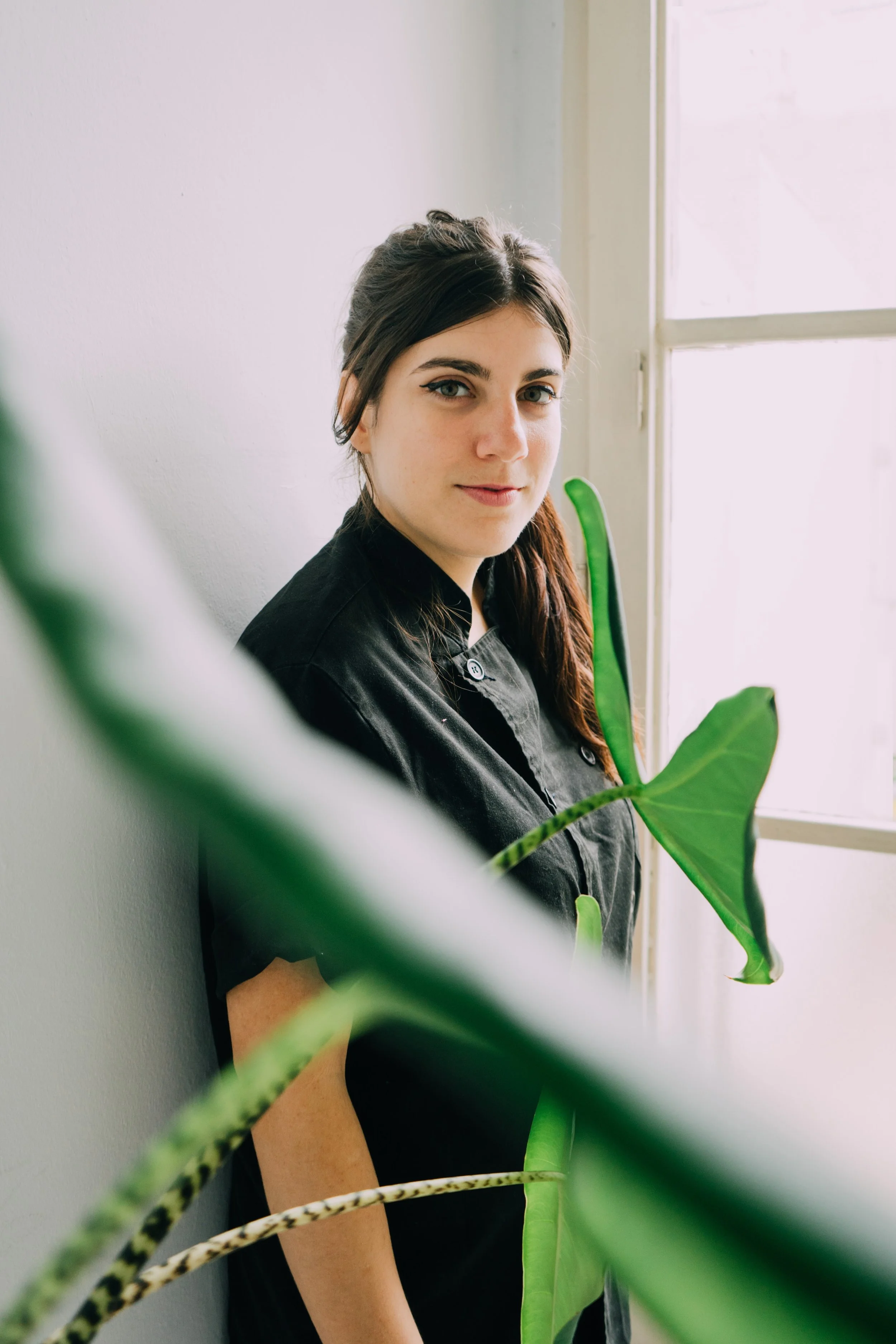 A young woman with long dark hair and fair skin, standing near a window and a green plant, looking at the camera with a slight smile.