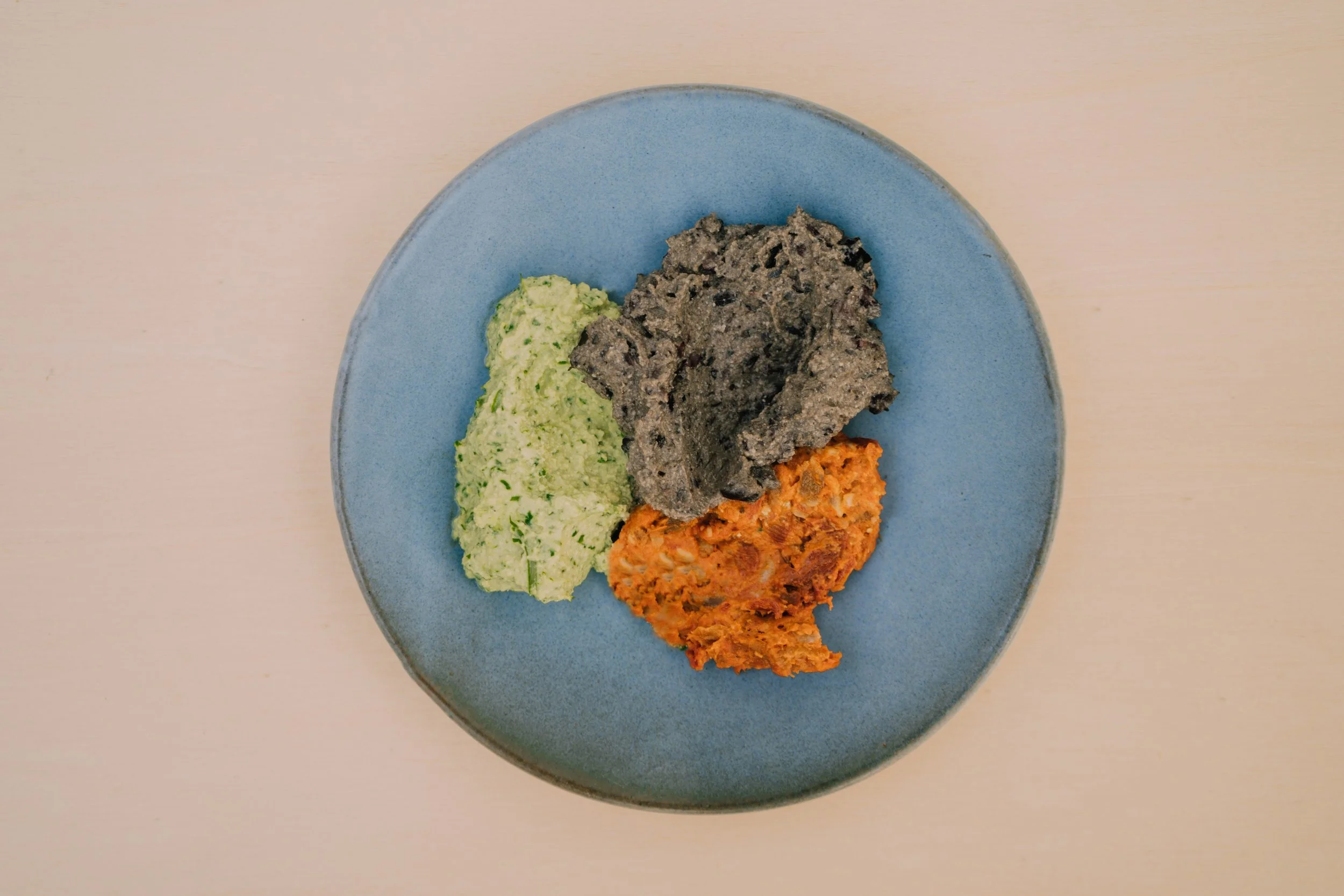 A blue plate with three different types of mashed vegetables or legumes, one grayish, one green, and one orange, on a light-colored wooden surface.