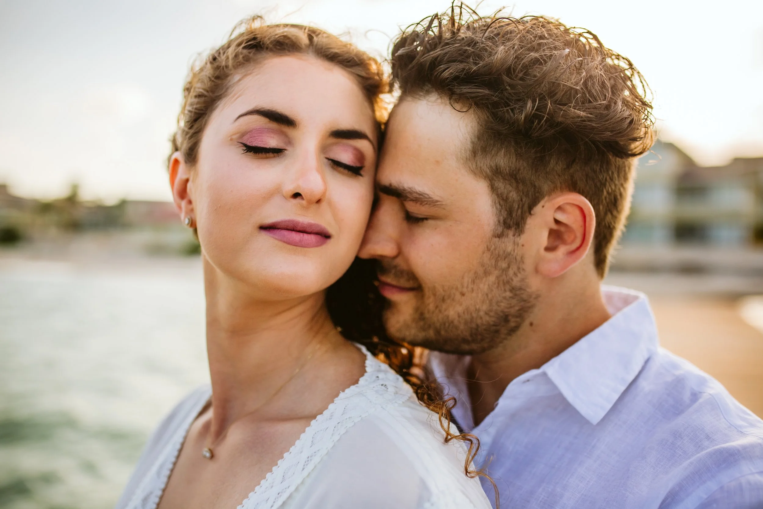 A close-up of a woman and a man, with their faces touching and eyes closed, outdoors near water during sunset.