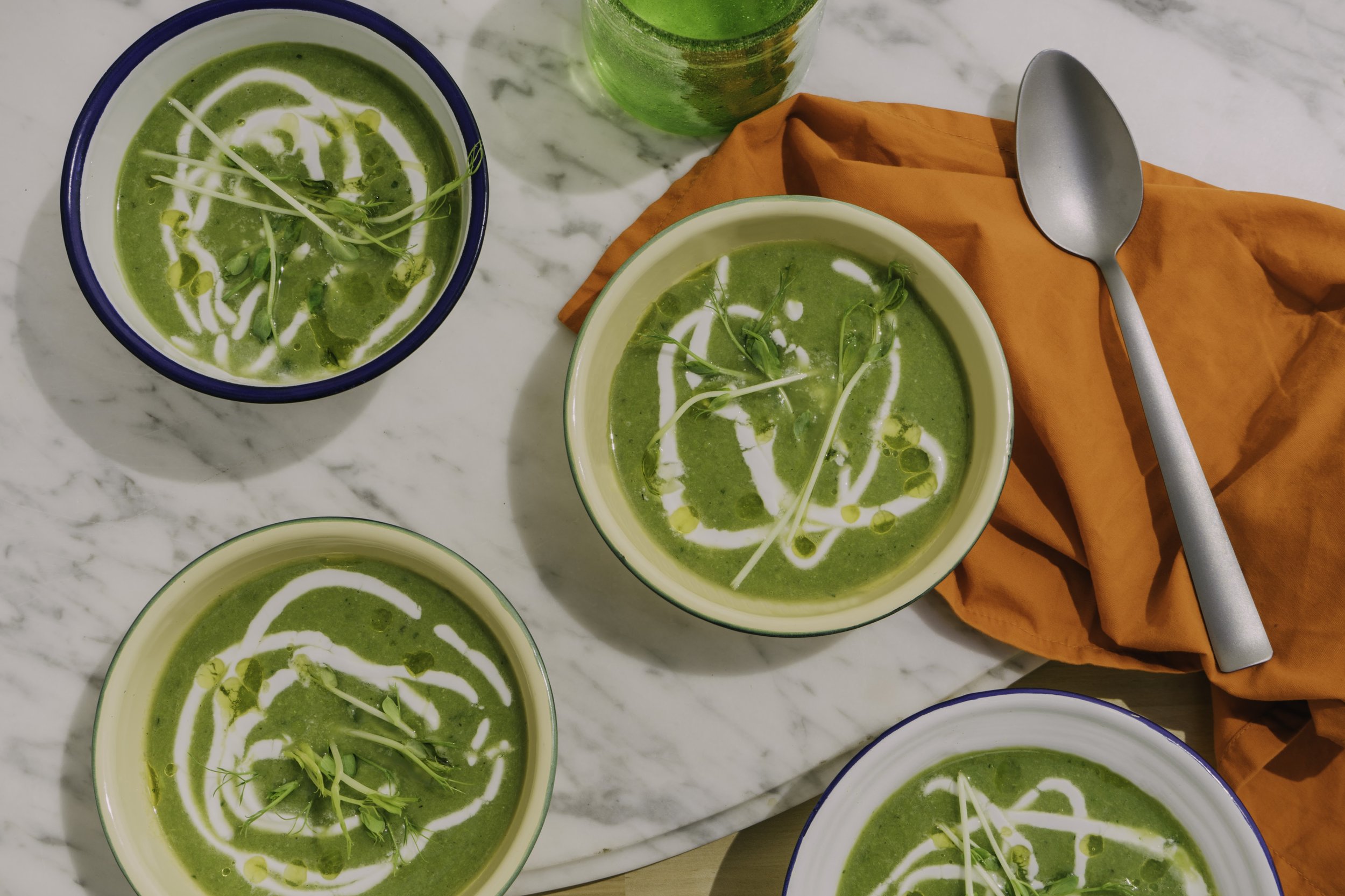 Four bowls of green soup garnished with microgreens and drizzled with cream, with a glass of green beverage, an orange napkin, and a silver spoon on a marble surface.