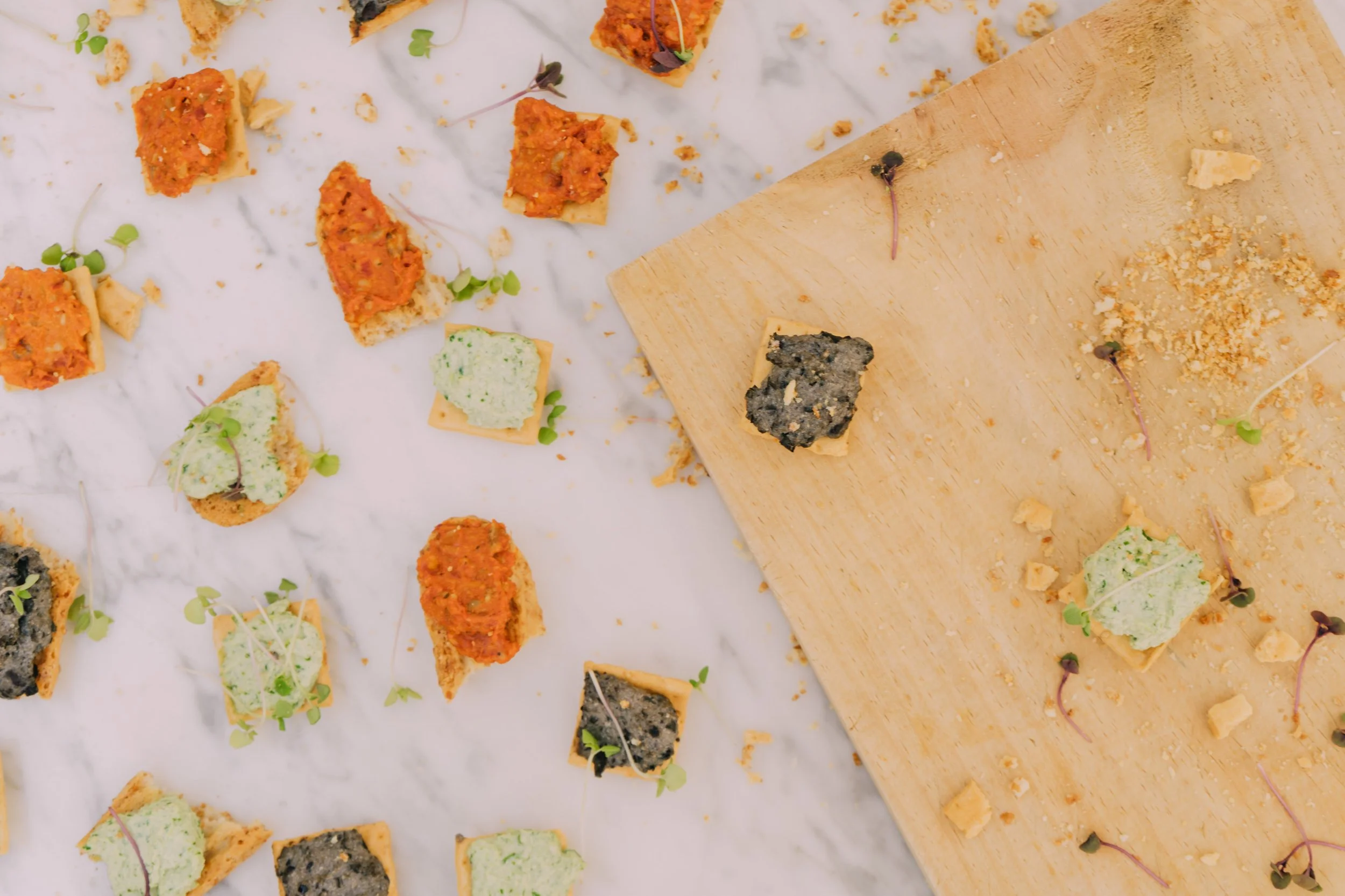 Assorted small appetizers on white parchment paper and a wooden board, including crackers with colorful spreads, garnished with microgreens.