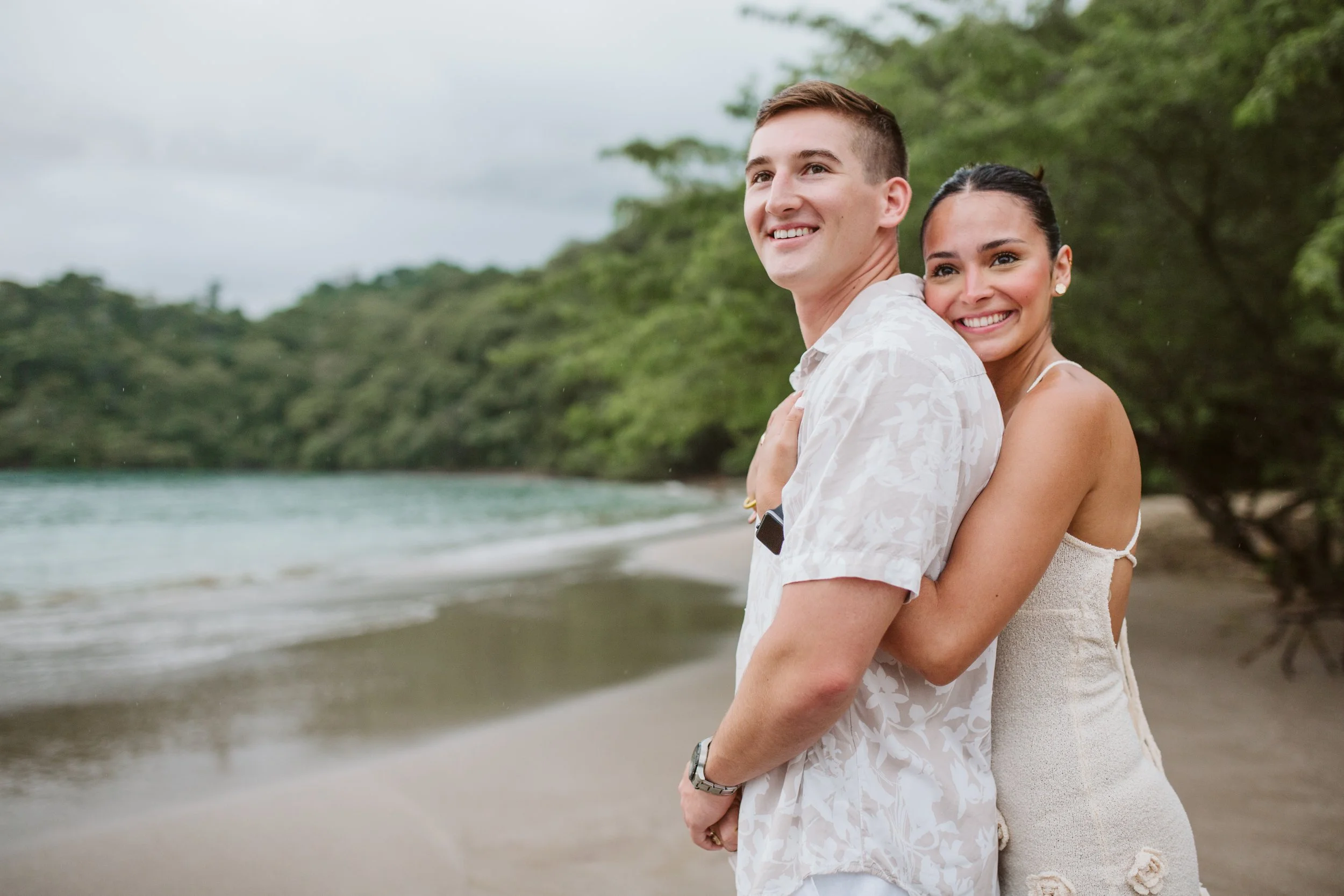 A happy couple stands on a beach, with the woman hugging from behind and smiling at the camera, while the man looks off into the distance, surrounded by trees and the ocean.