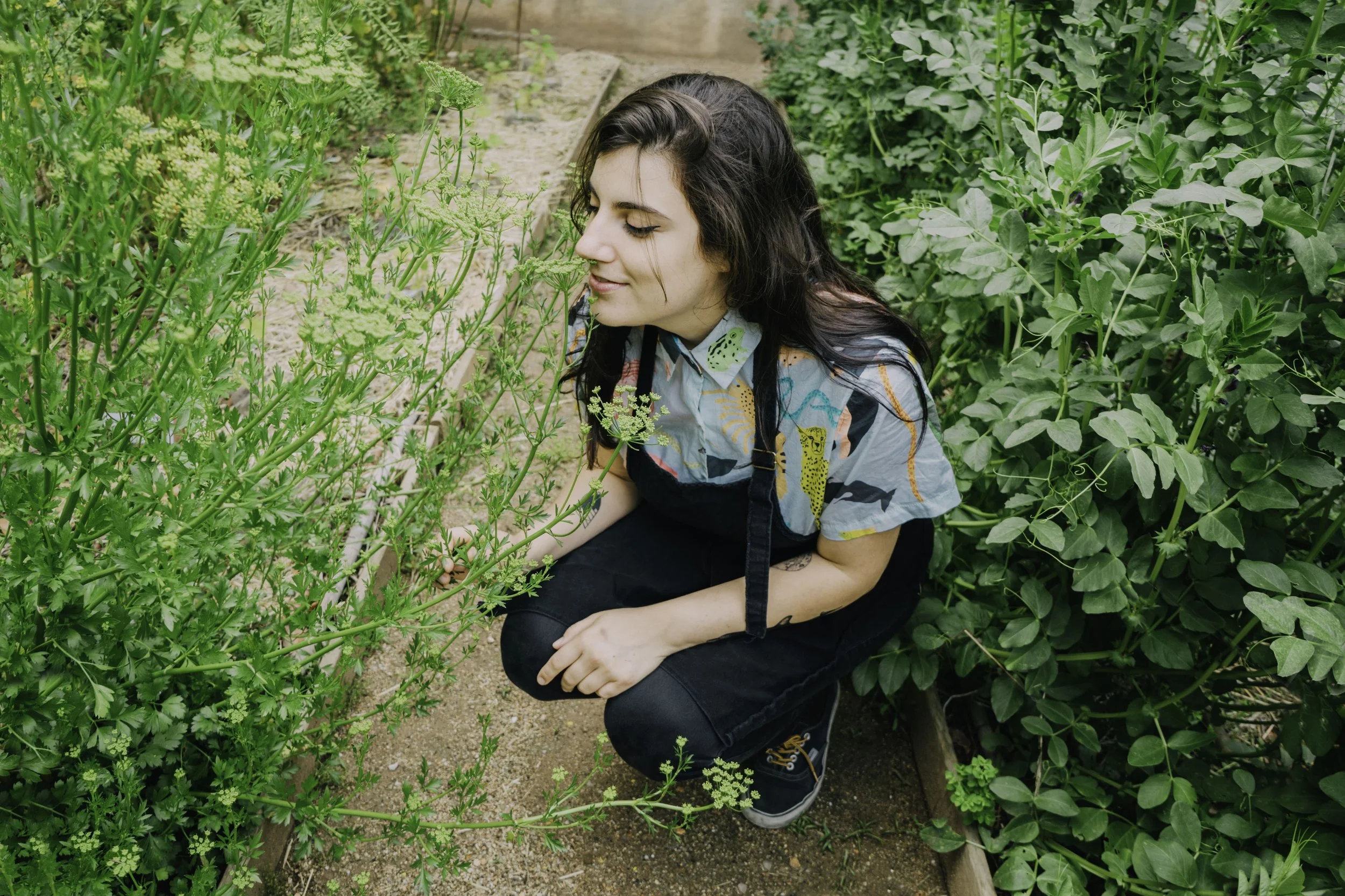 Woman kneeling in a garden among green plants, smelling a flowering plant.