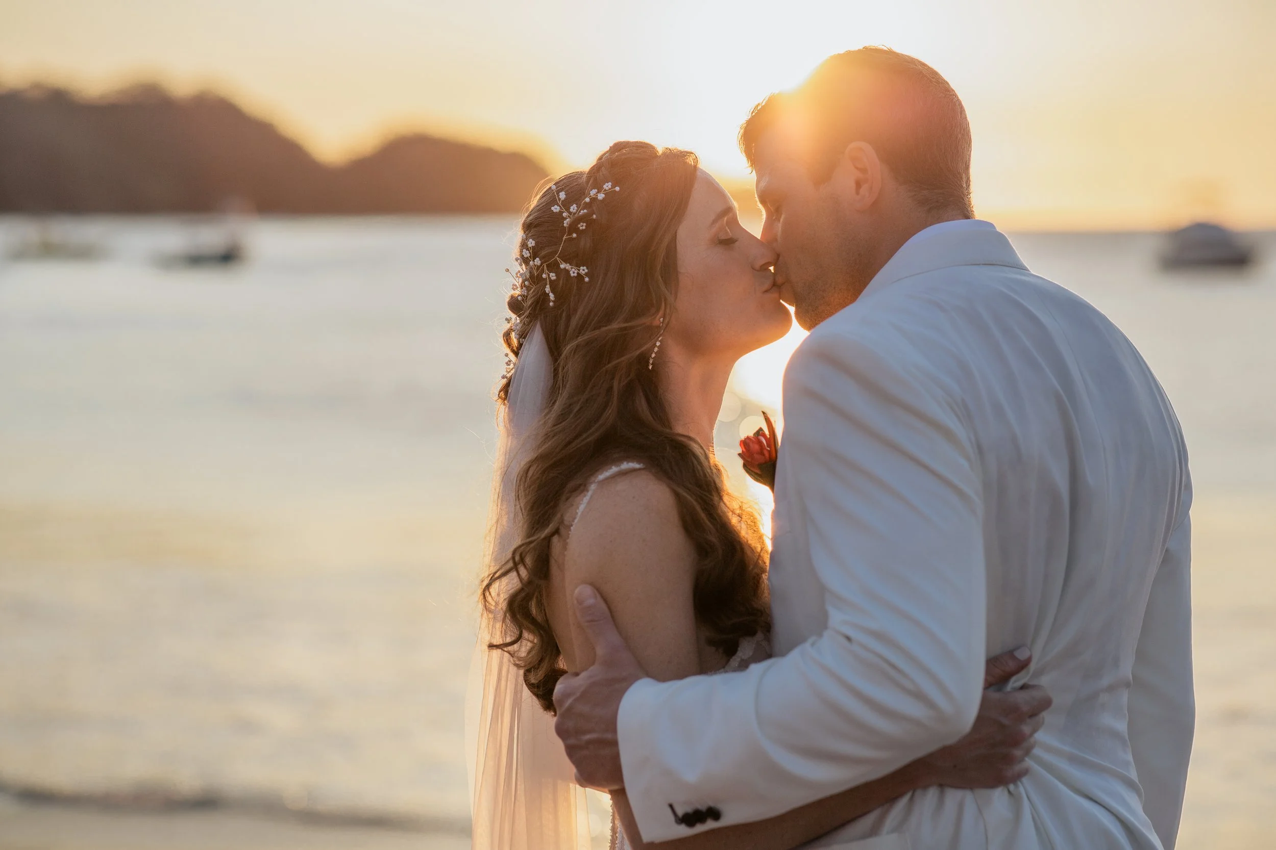A couple kissing on the beach during sunset, with boats in the water and mountains in the background.