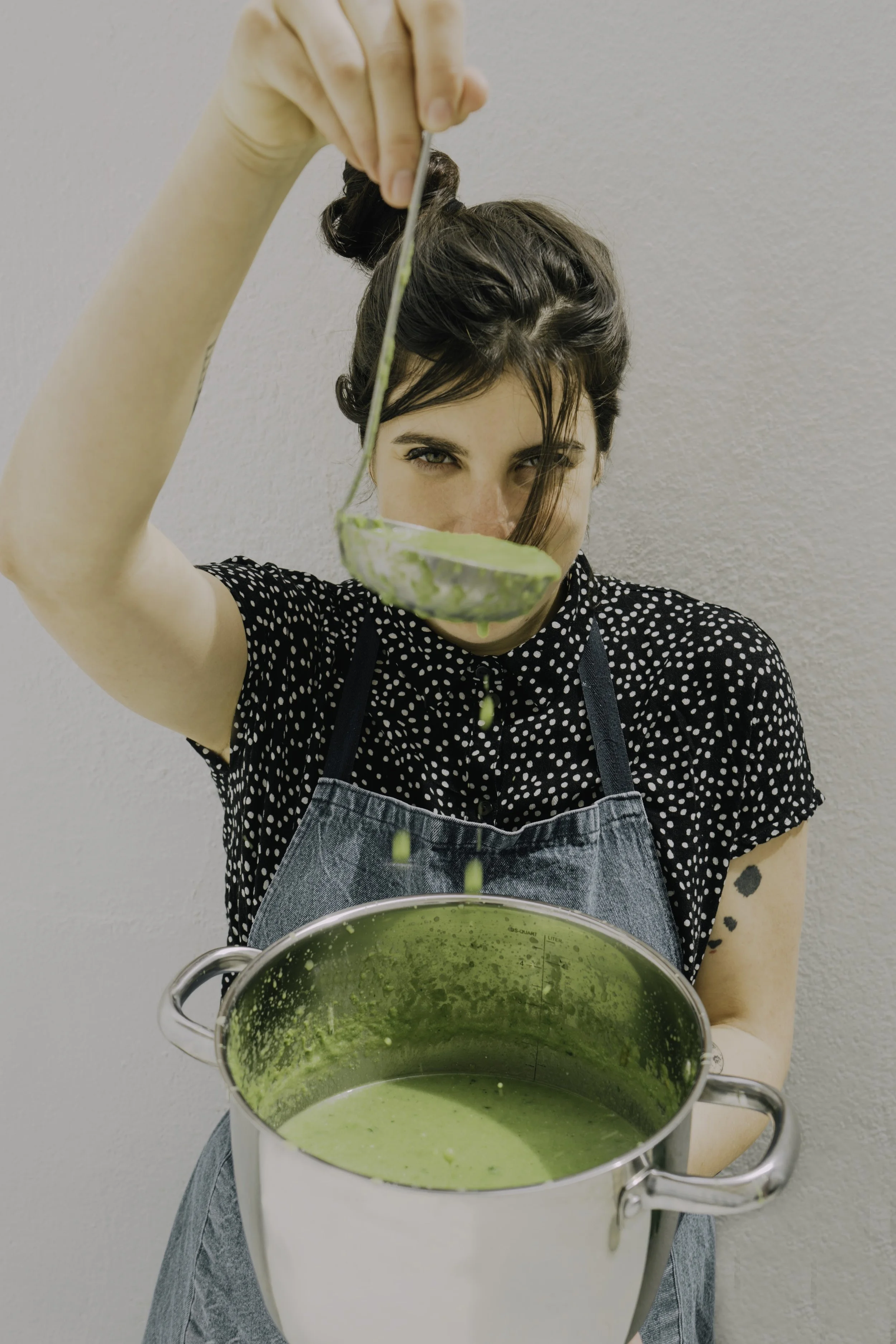 Woman in black polka dot shirt and apron holding a ladle with green soup, standing in front of a plain white wall.