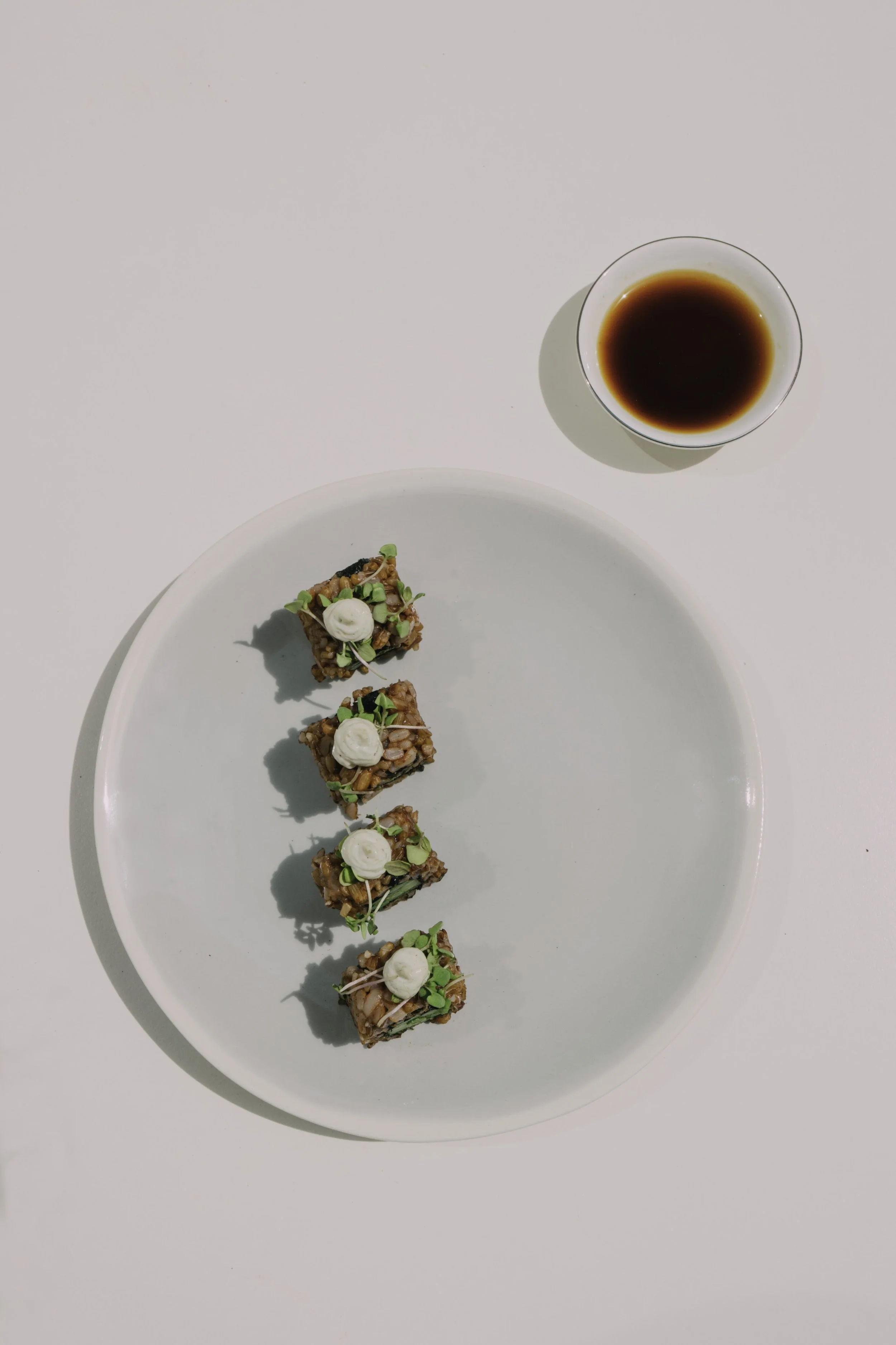 A white plate with four small pieces of sushi topped with cream and green garnishes, a bowl of soy sauce, and a white background.