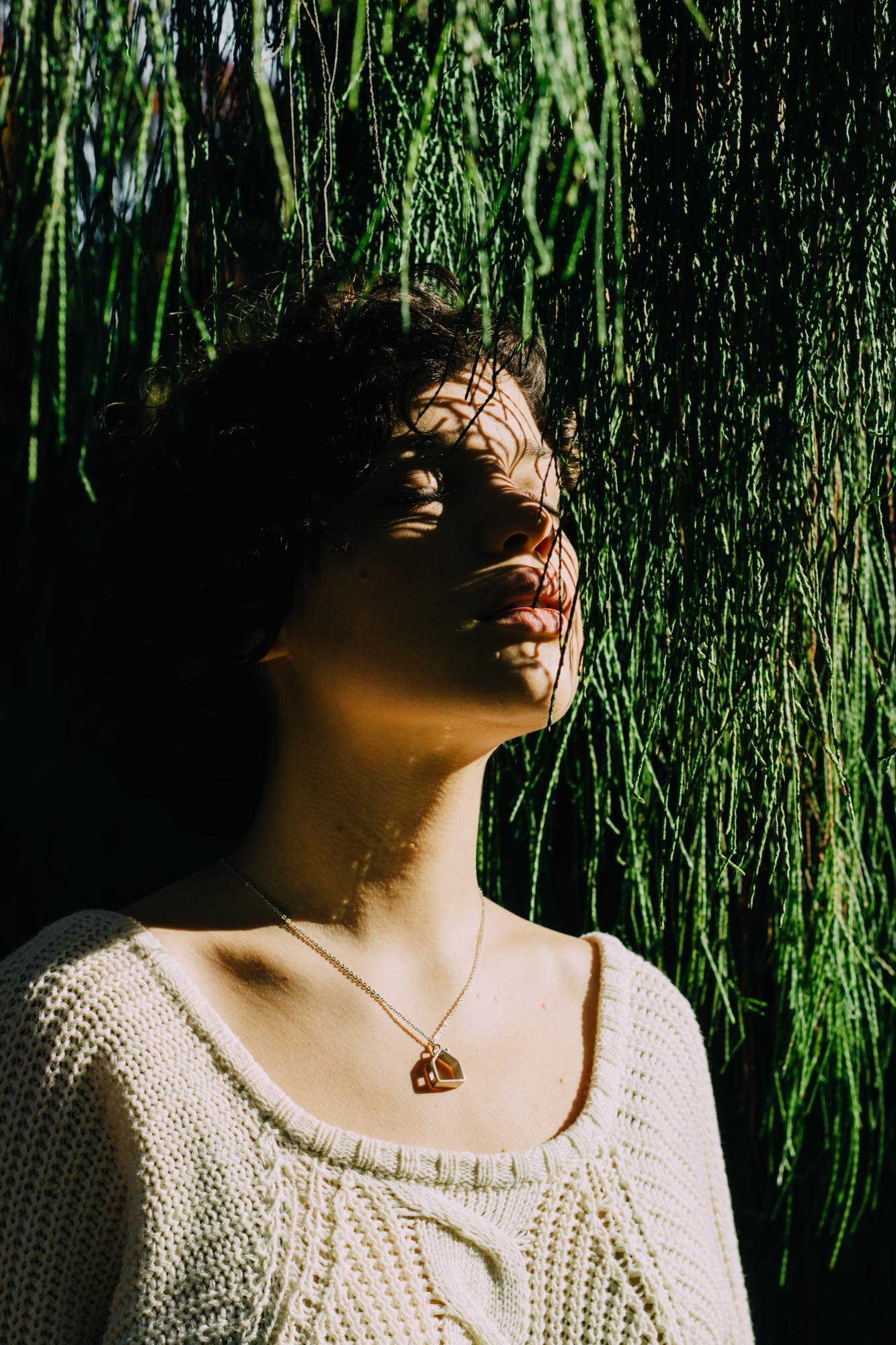 A woman with curly dark hair standing in front of green bamboo stalks, casting shadows on her face from the sunlight.