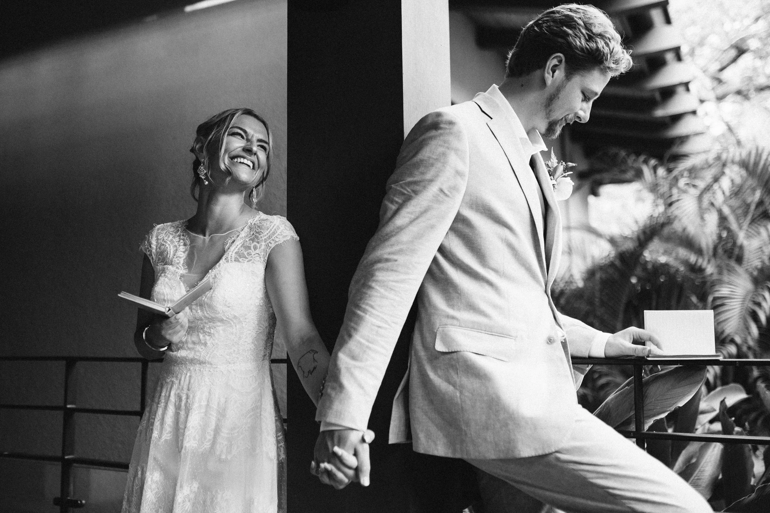 A black and white photo of a happy bride and groom holding hands, with the bride smiling and looking at the groom who is reading from a book.