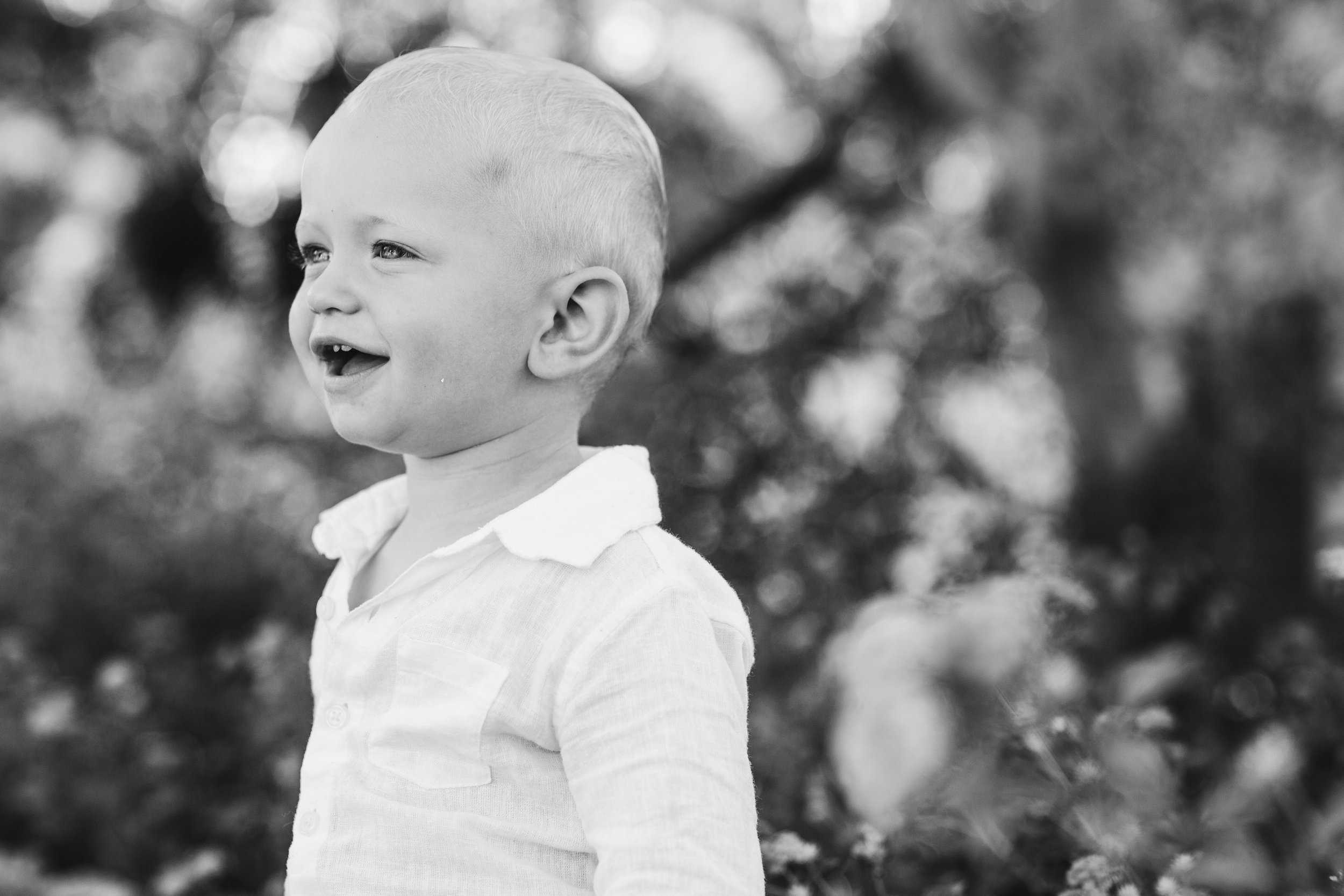 A young boy with short light-colored hair, smiling and looking to the side outdoors, dressed in a light-colored button-up shirt, with blurred trees and foliage in the background.