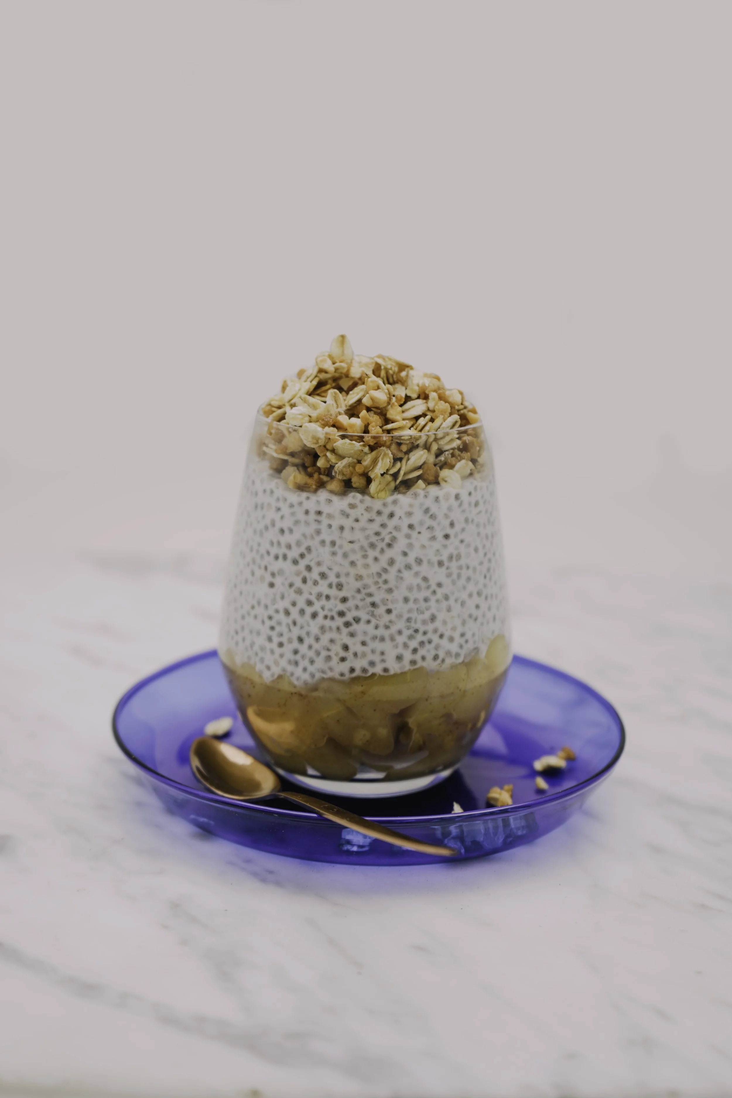 A glass of chia seed pudding topped with granola, served on a blue plate with a small spoon and some granola scattered around, on a white marble surface.