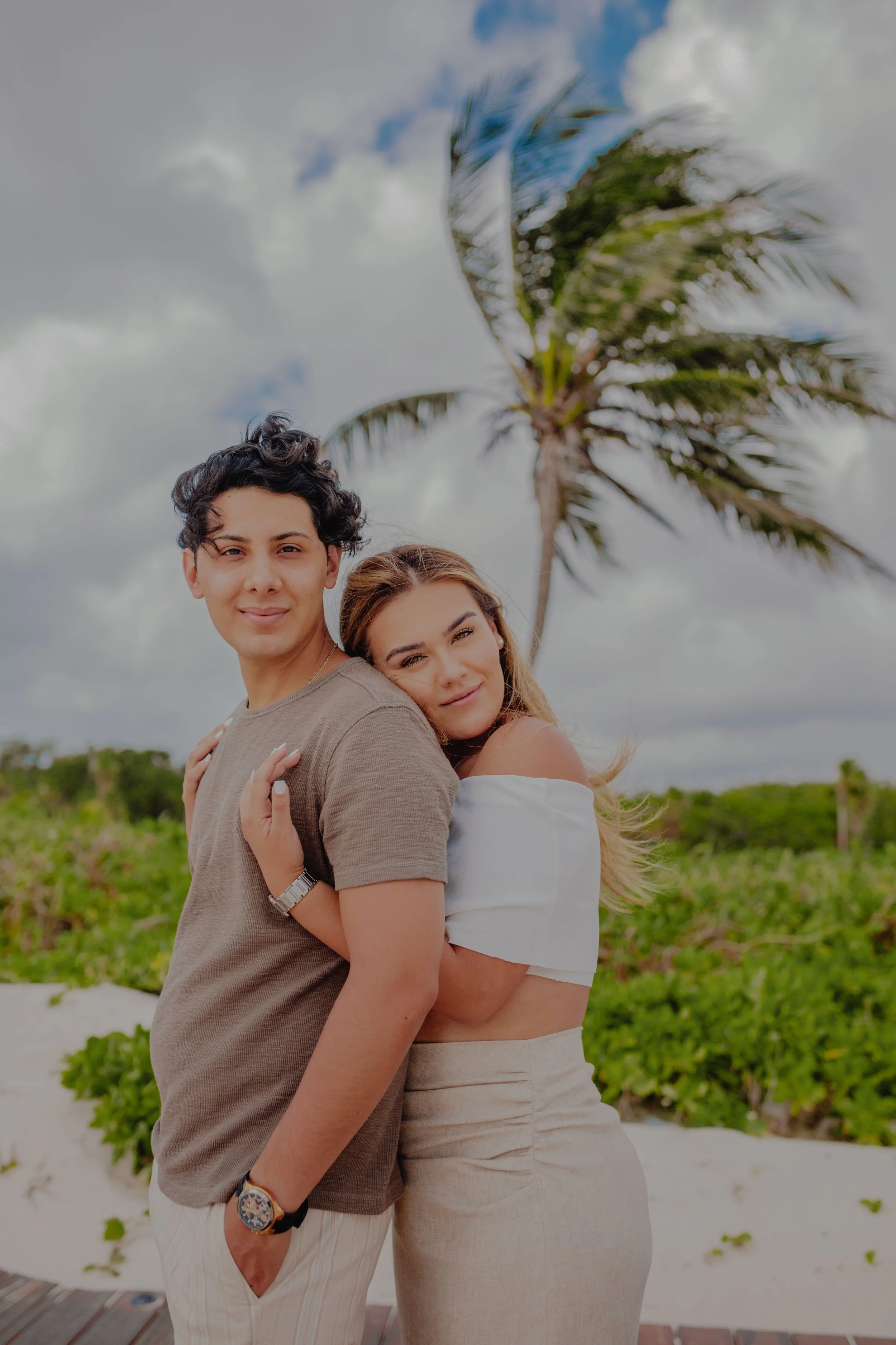 A young couple at the beach, standing close together with a palm tree and cloudy sky in the background.