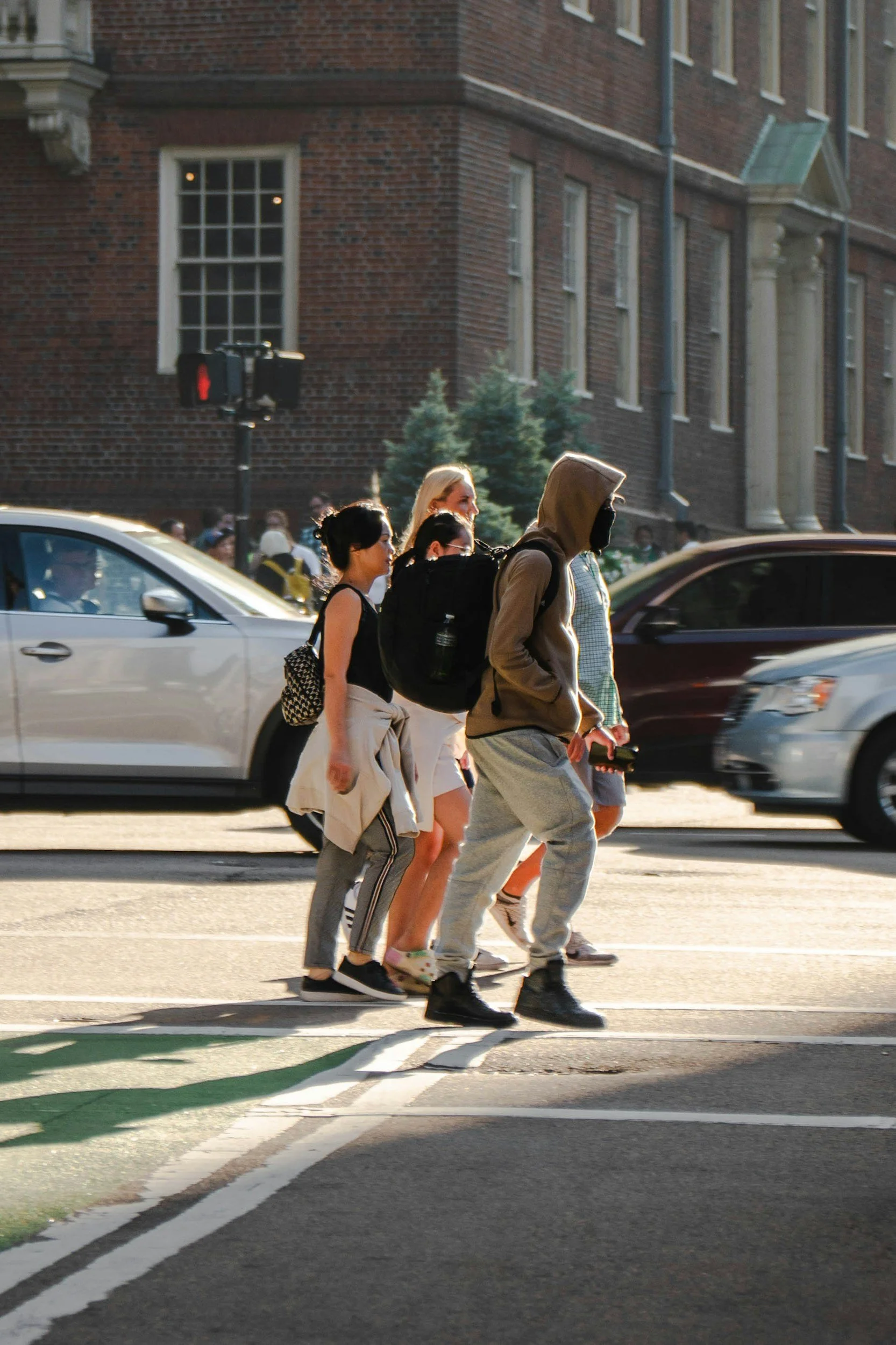 A group of five pedestrians crossing the street at a crosswalk in an urban area, with cars passing by and brick buildings in the background.