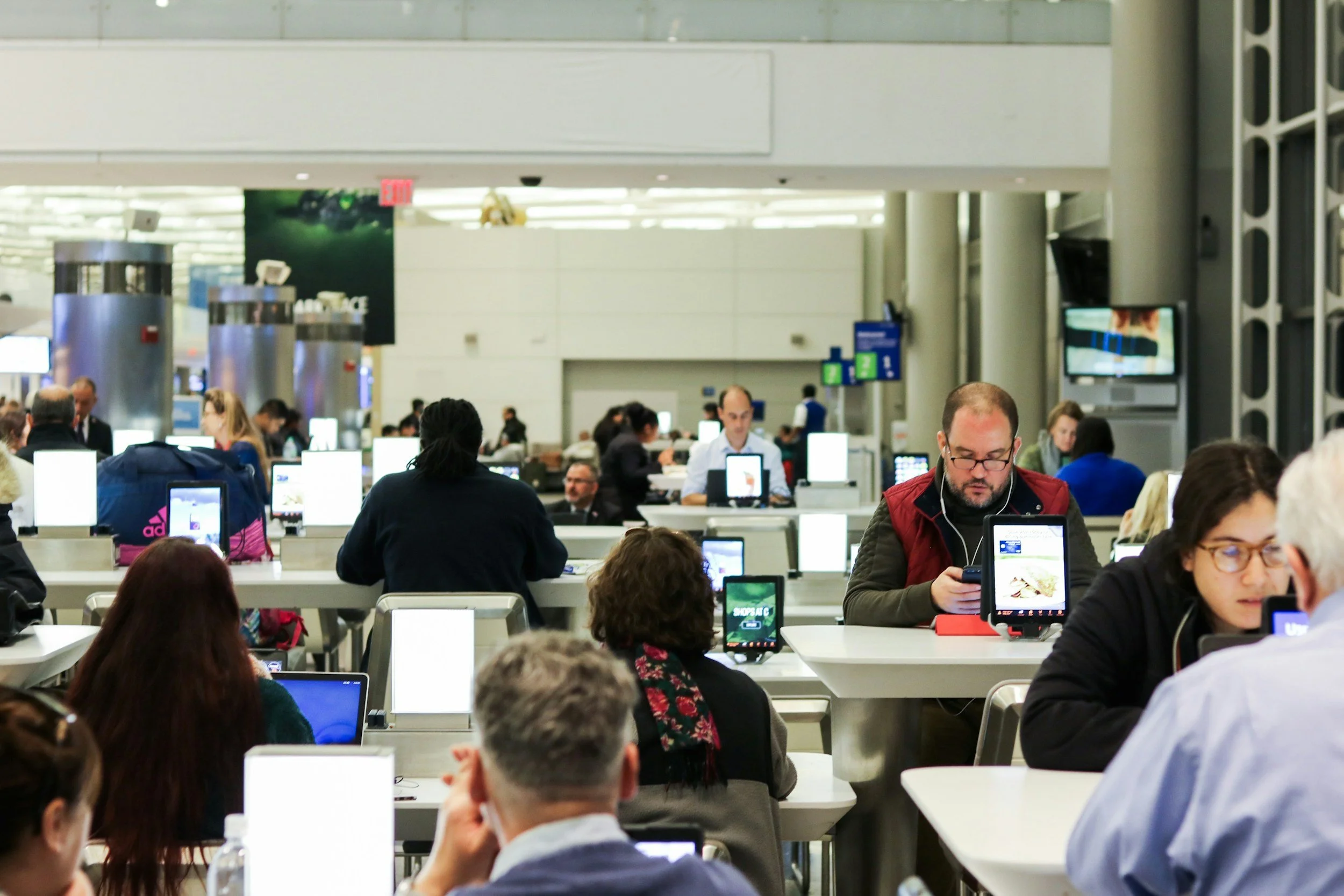 People sitting at tables in an airport food court, using electronic devices and waiting.